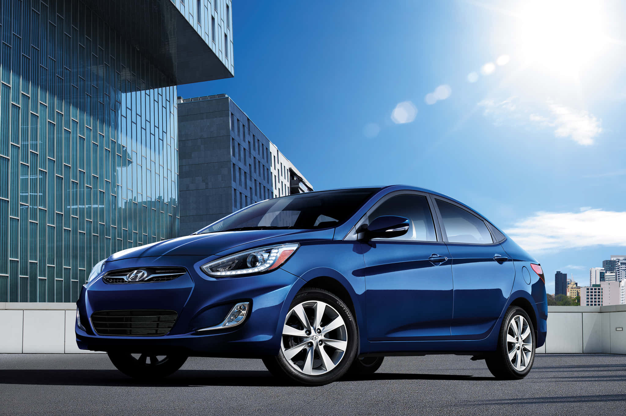 Hyundai Accent 桌布 壁紙 — 一輛時尚的藍色現代轎車在明亮的陽光下閃耀，背景是現代化的屋頂和玻璃摩天大樓。