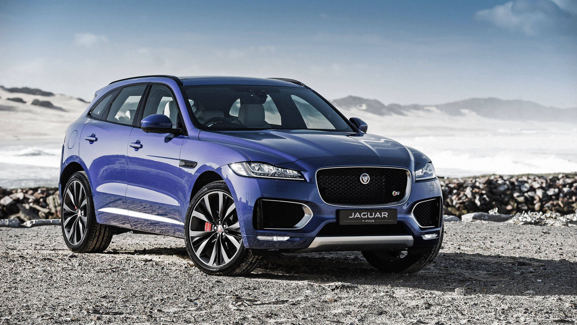 藍色Jaguar F-Pace SUV停泊在靠近雪山的岩石海灘上。 — 美洲豹汽車 壁紙