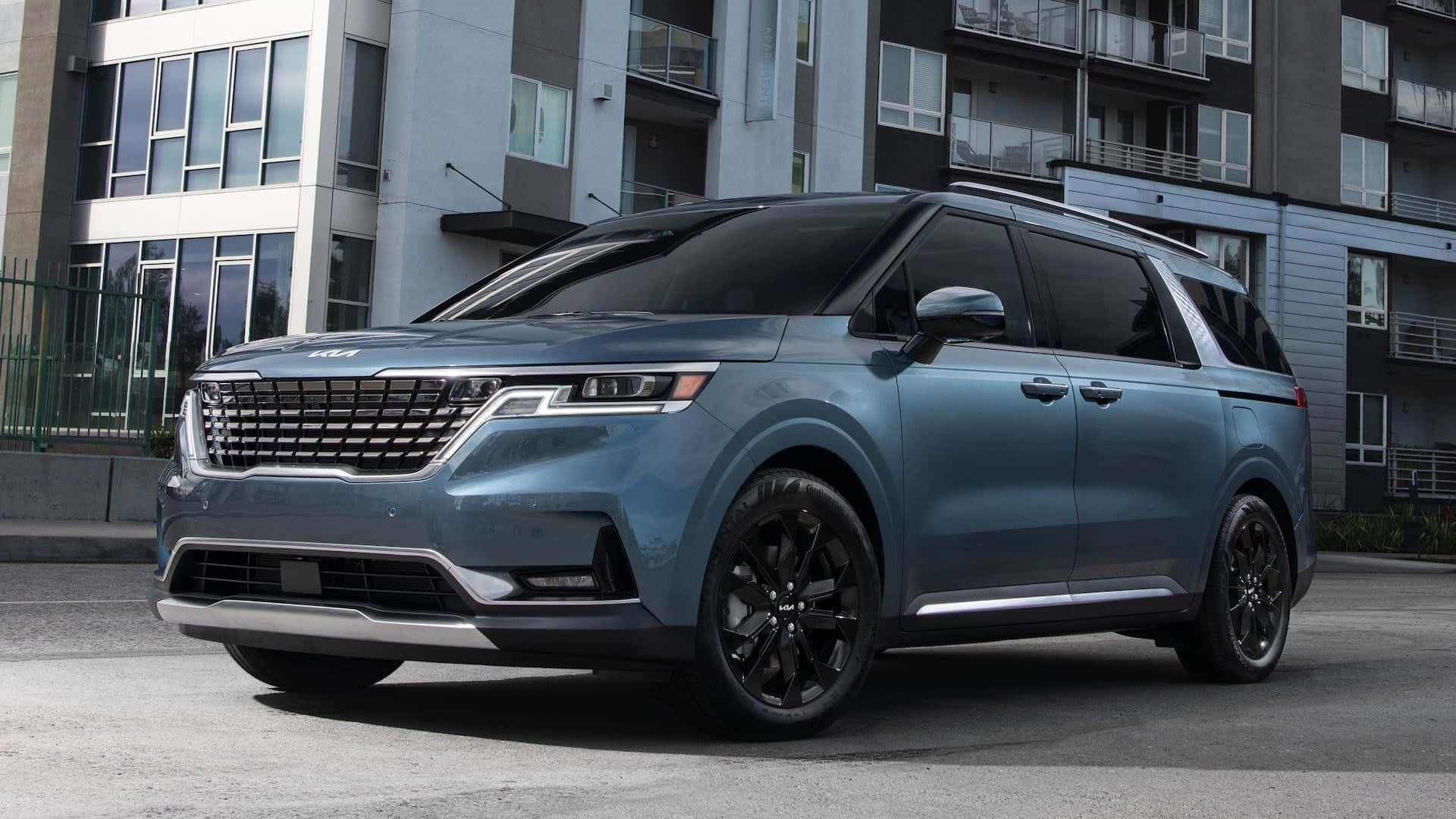 Kia Carnival 桌布 壁紙 — 一架時尚的藍色Kia小型貨車配黑色輪圈，停泊在現代公寓大樓前的城市街道上。