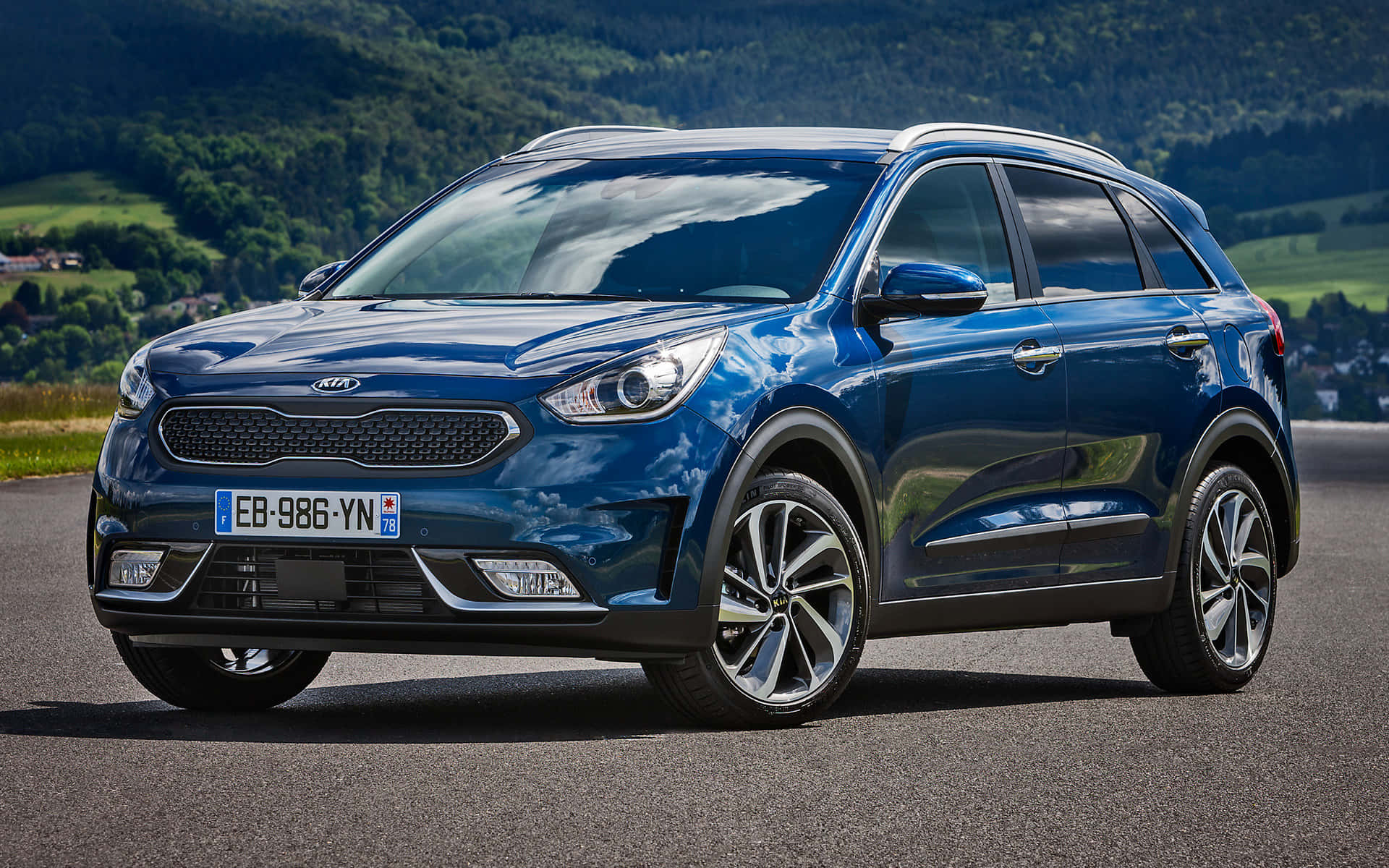 Kia Niro 桌布 壁紙 — 一輛時尚的藍色Kia SUV停在柏油路上，背景是起伏的綠色山丘和樹木，在明亮的日光下。