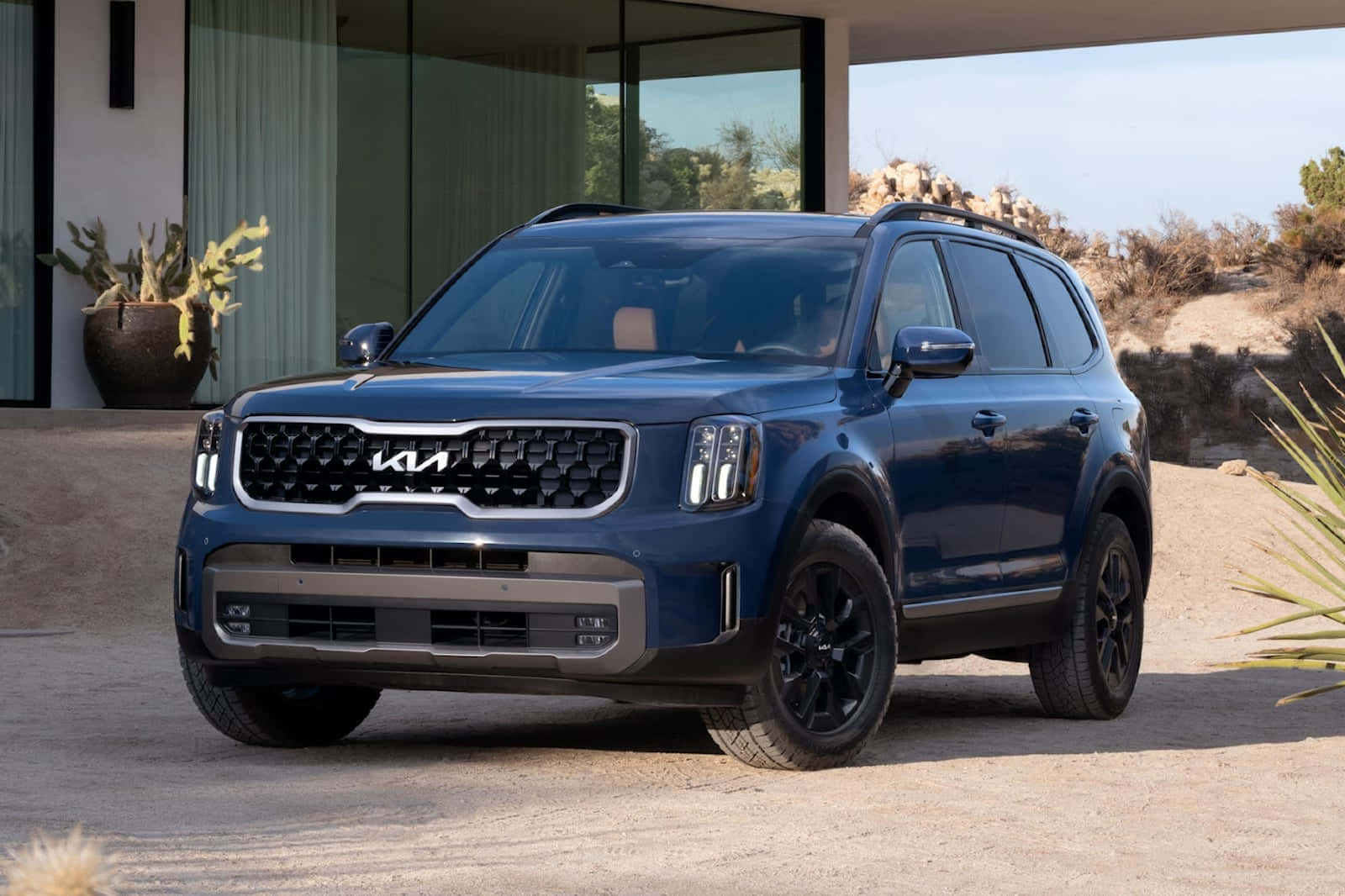 Kia Telluride 桌布 壁紙 — 一架時尚的藍色Kia SUV停泊在沙漠車道上，旁邊是帶玻璃牆和仙人掌的現代房屋。