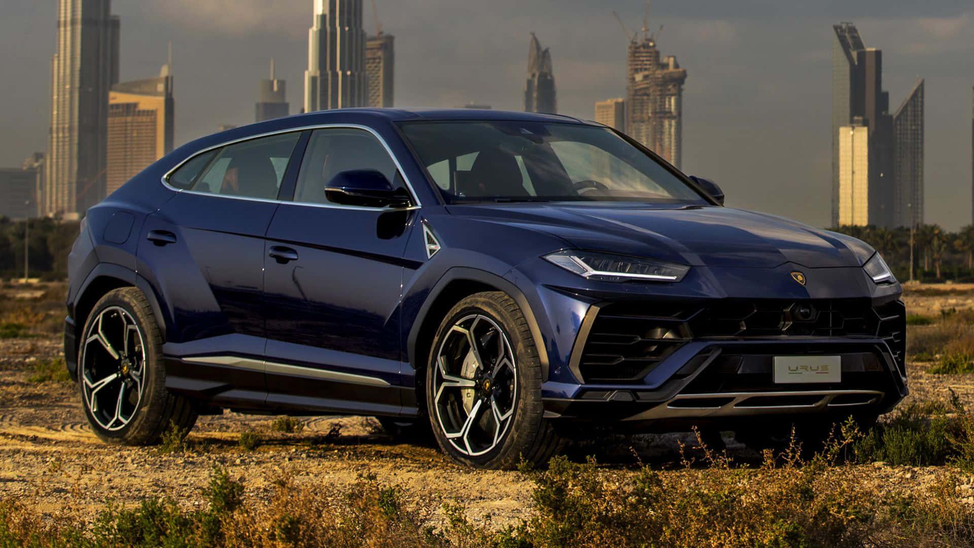 Lamborghini Urus 桌布 壁紙 — 一架時尚的藍色藍寶堅尼 Urus SUV 立於乾草上，背景是現代城市天際線。