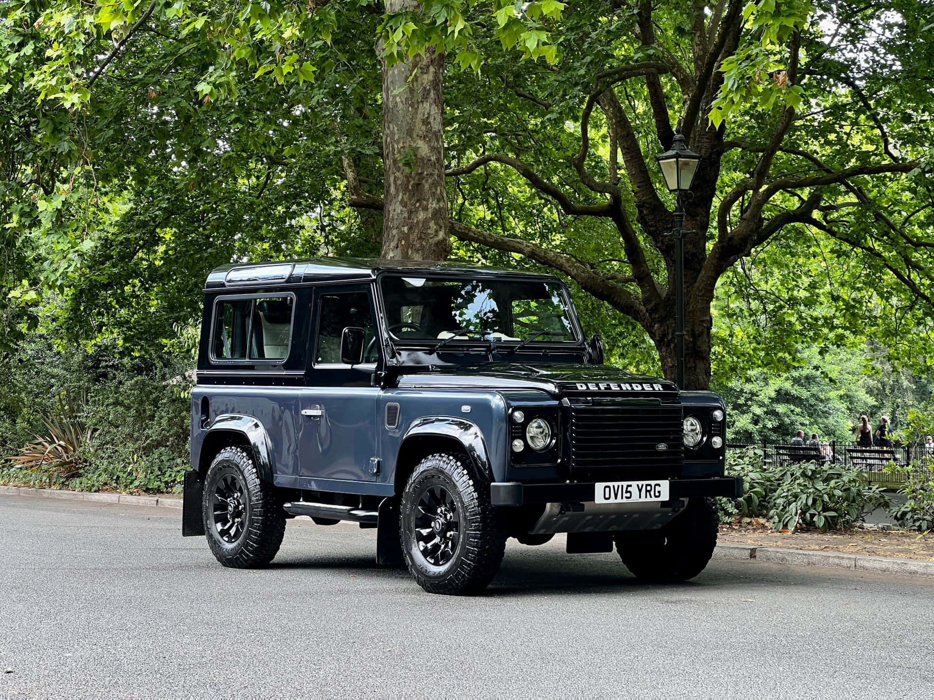 Land Rover Defender 桌布 壁紙 — 一輛時尚的藍色 Land Rover Defender 停放在陽光普照、綠樹成蔭的公園道路上。