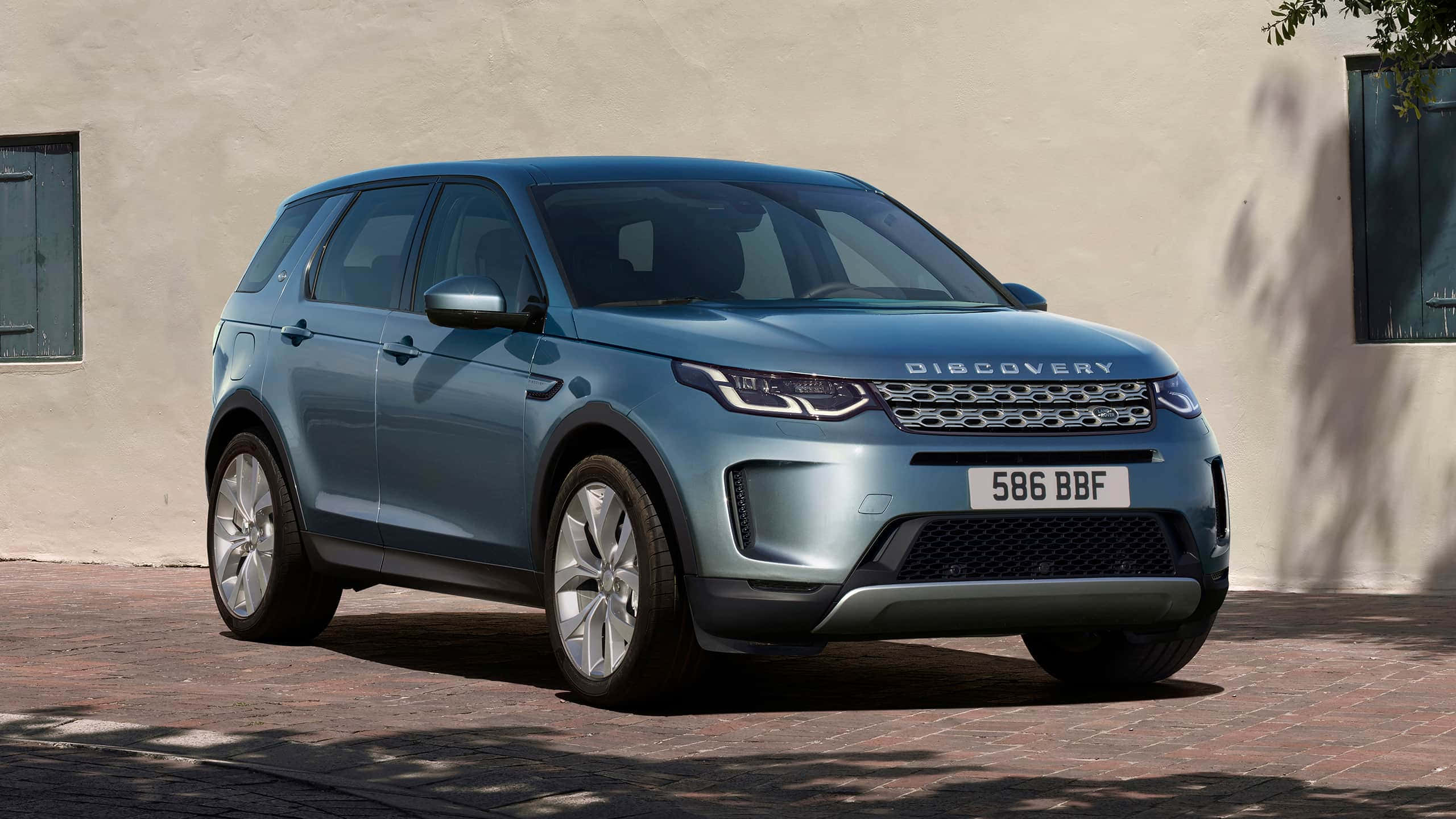 Land Rover Discovery 桌布 壁紙 — 一輛時尚的藍色 Land Rover Discovery 停放在陽光明媚的磚砌車道上，旁邊是帶綠色百葉窗的米色灰泥牆。