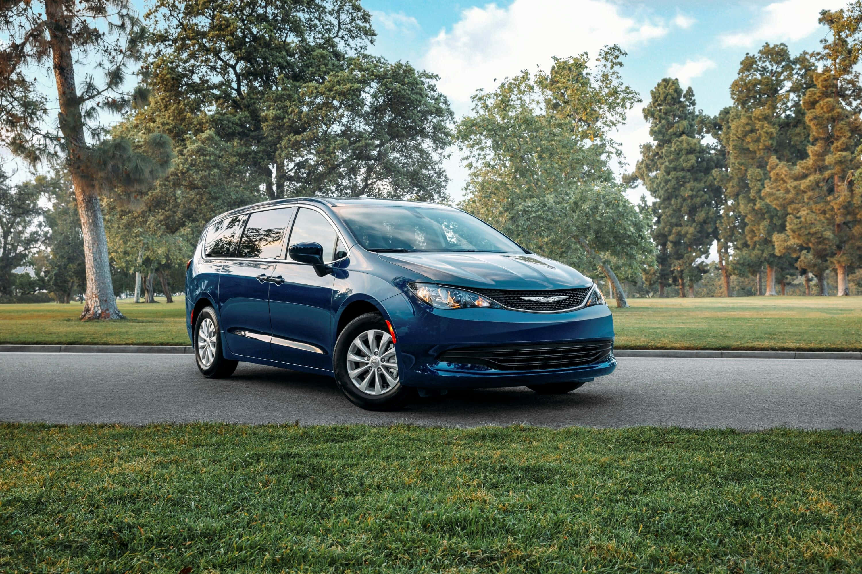 Chrysler Voyager 桌布 壁紙 — 一架時尚的藍色小型貨車停泊在鋪砌的道路上，周圍是茂密的綠草和高大的樹木，天空明亮。