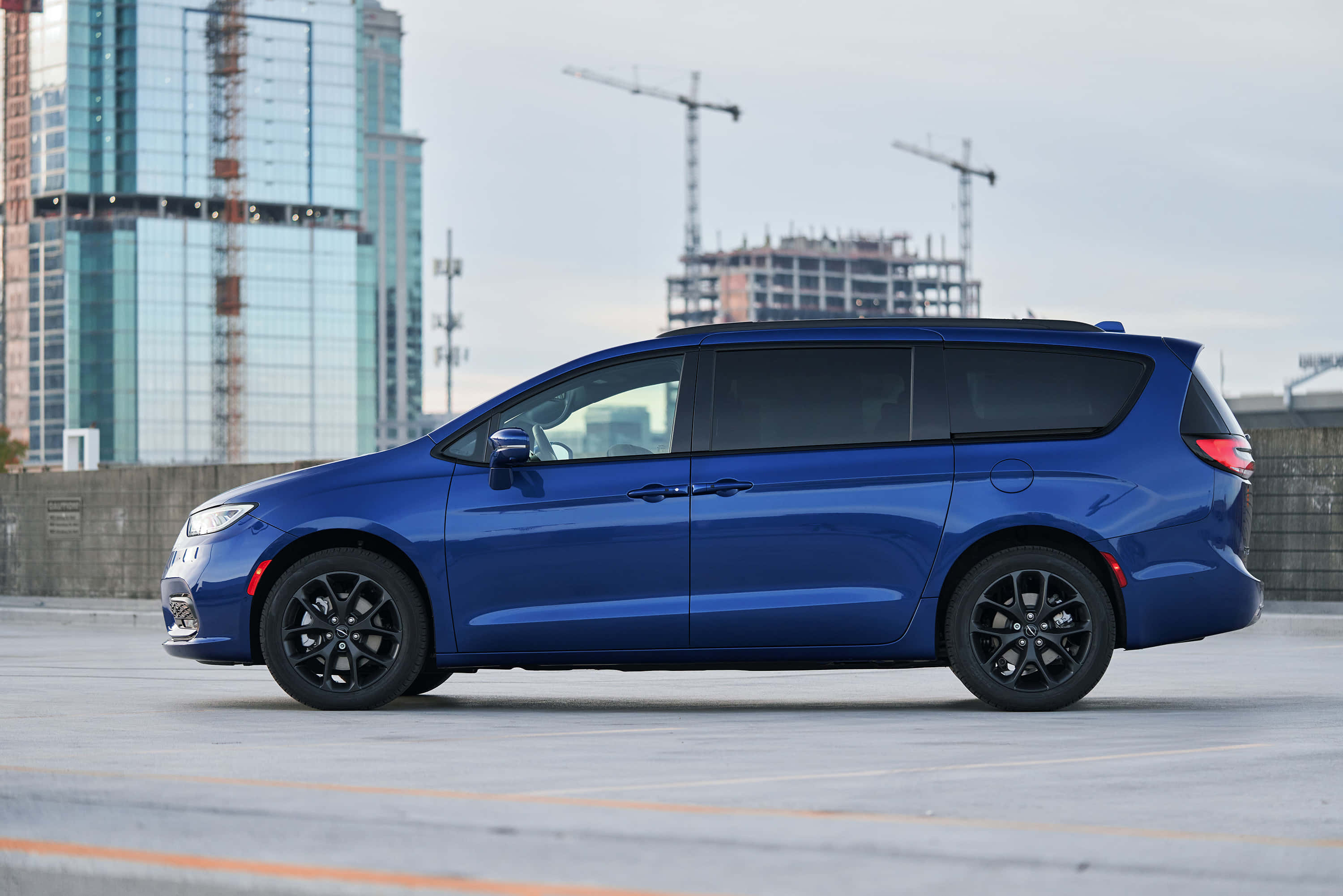 Chrysler Pacifica 桌布 壁紙 — 一架時尚的藍色小型貨車配黑色輪圈，停放在城市空地，背景是玻璃摩天大樓和建築起重機。