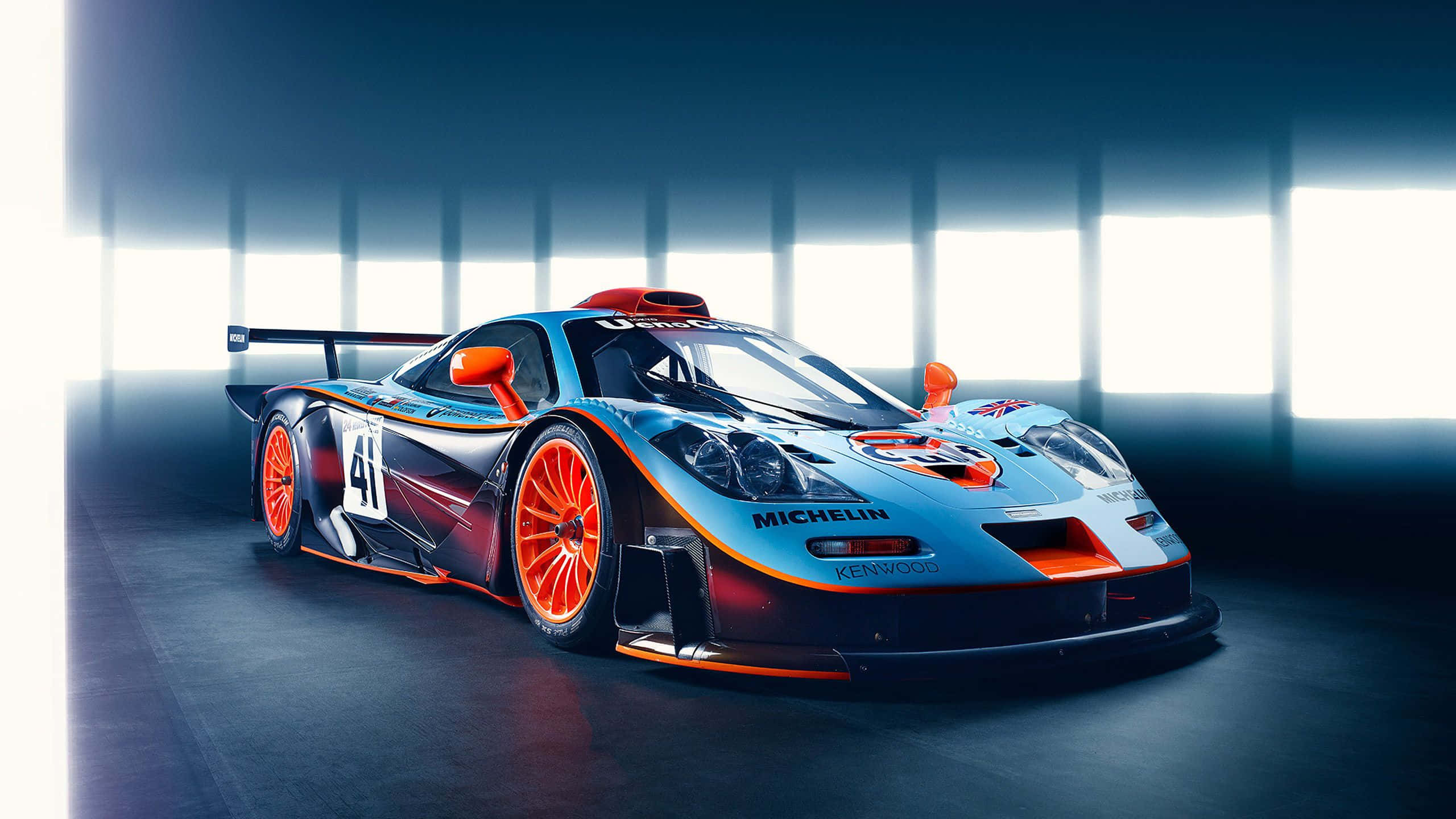 Mclaren F1 桌布 壁紙 — 一輛時尚的藍色和橙色賽車，號碼41，展示在光線昏暗的車庫中，背後有明亮的垂直窗戶。