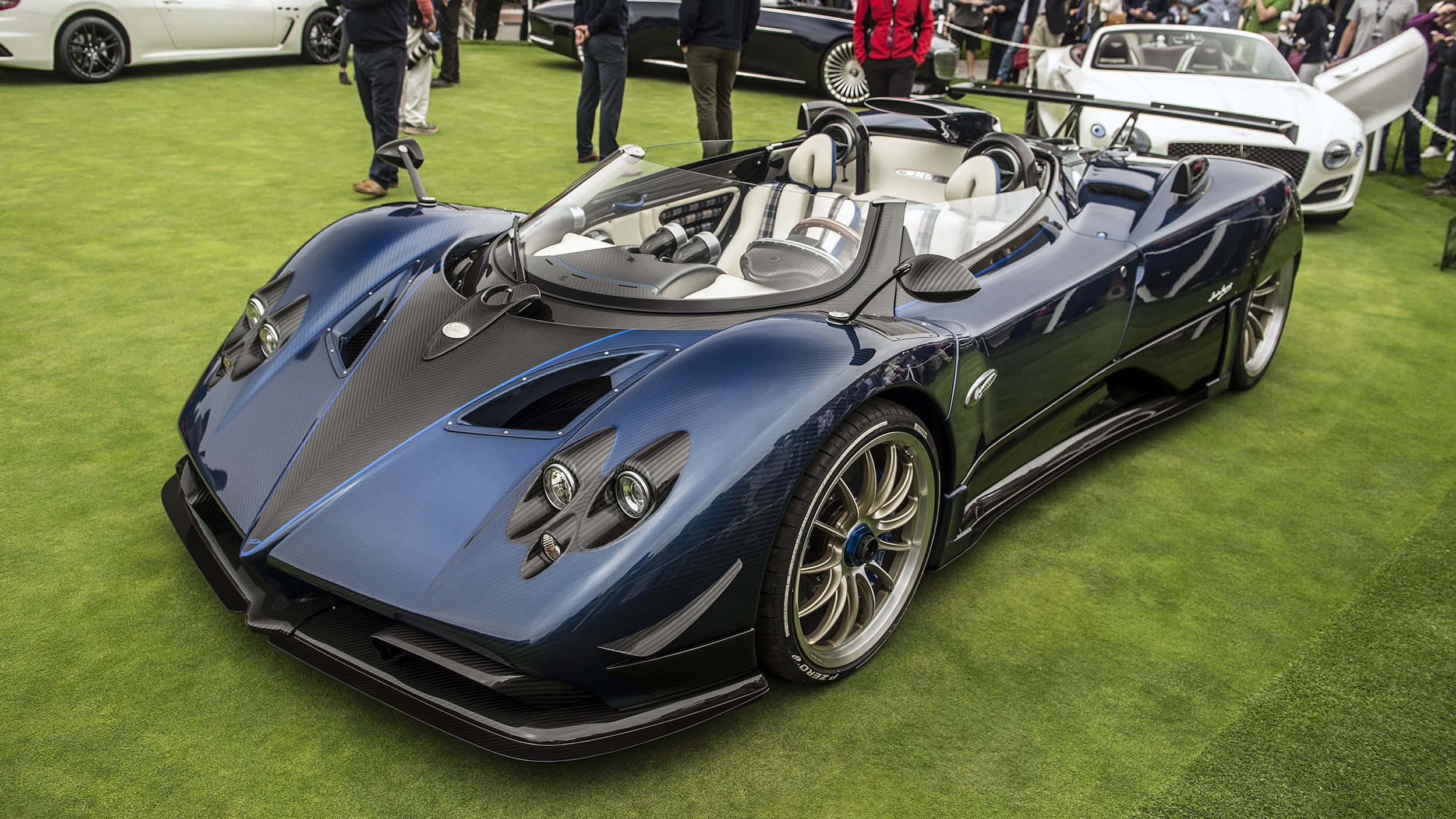 Pagani Zonda Hp Barchetta 桌布 壁紙 — 一輛流線型的藍色帕加尼 Huayra 敞篷跑車在車展的綠色地毯上閃耀著明亮燈光，人們正在欣賞它。