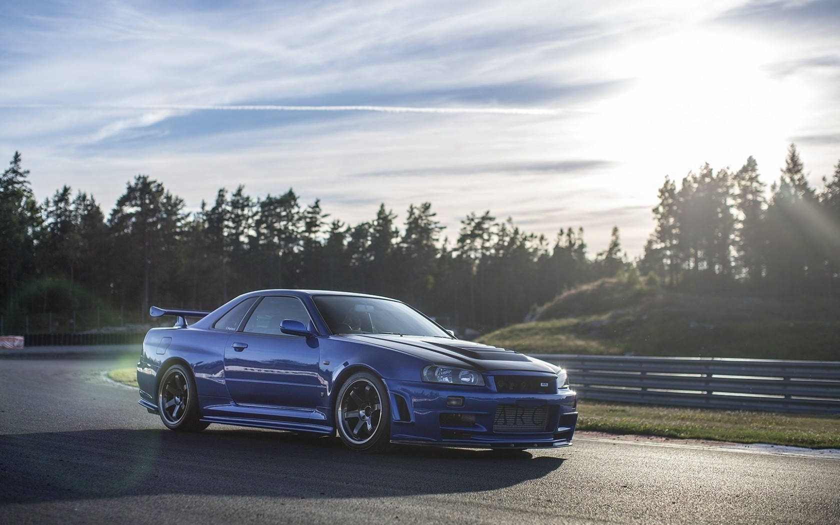 藍色跑車在賽道上漂移，周圍有松樹和夕陽餘暉。 — 日產 Skyline GTR R34 壁紙