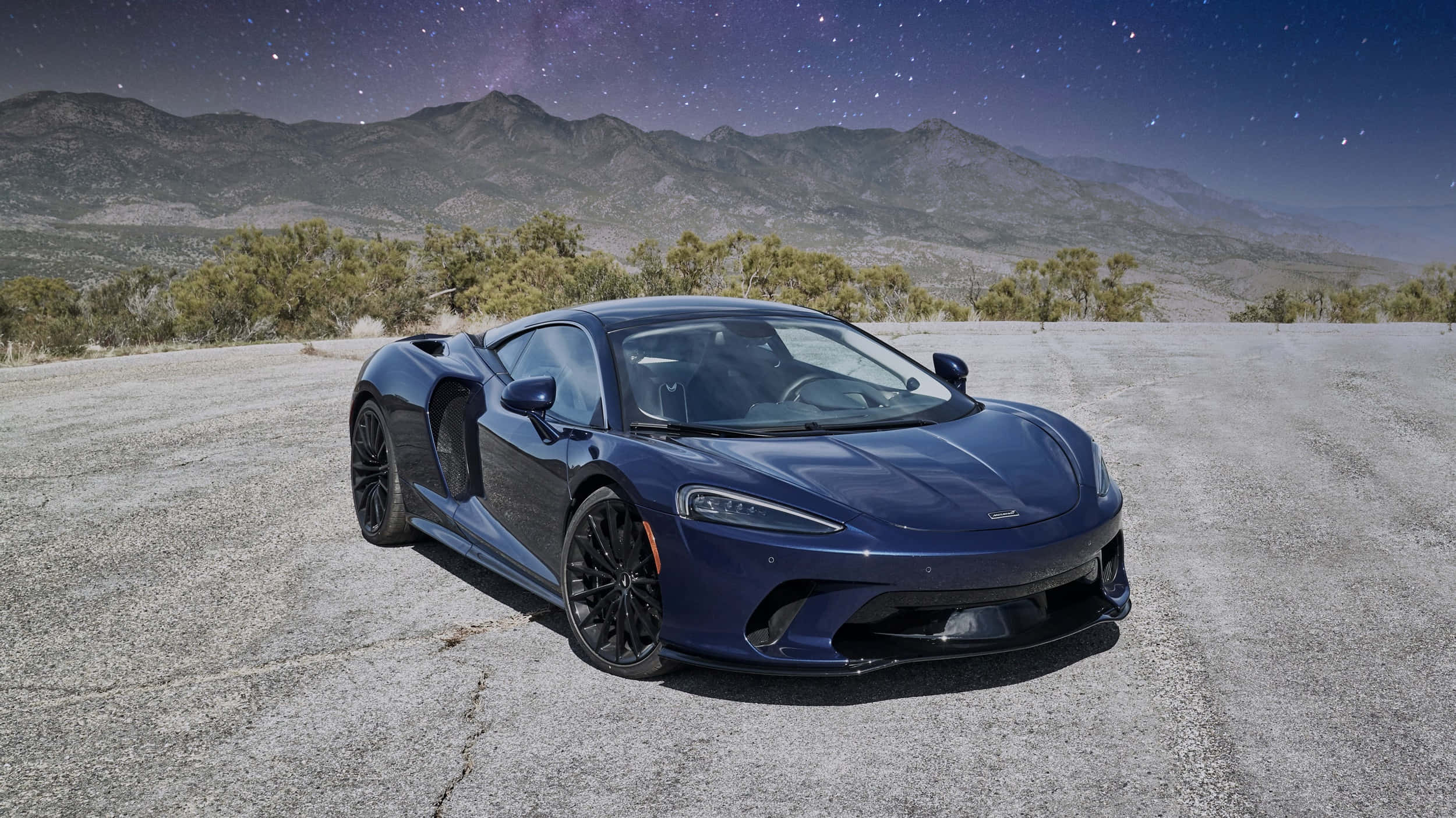 Mclaren Gt 桌布 壁紙 — 一輛時尚的藍色跑車停在裂開的路面上，背景是星空和山脈。