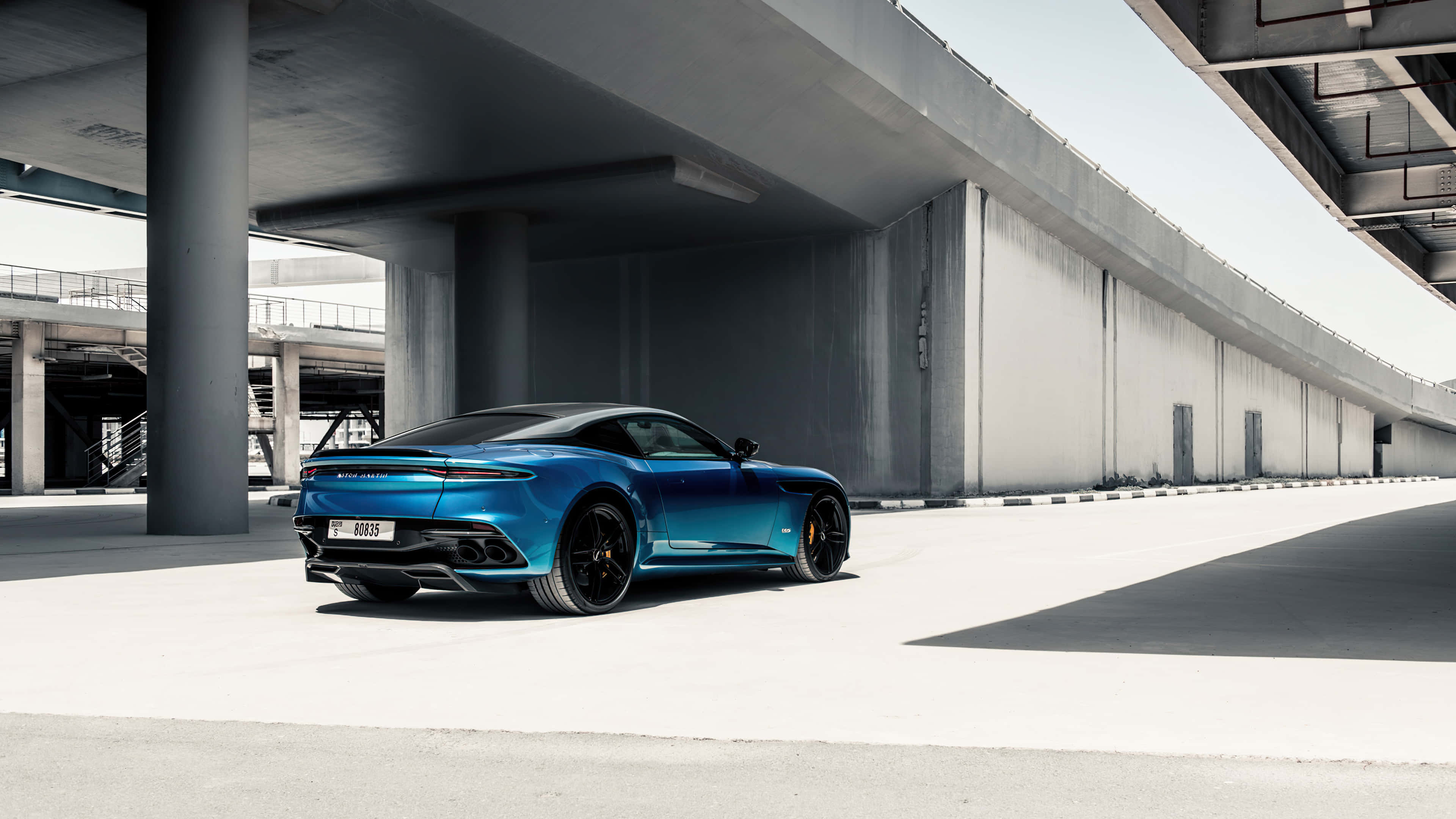 Aston Martin Dbs Superleggera 桌布 壁紙 — 一架時尚的藍色跑車停放在現代混凝土天橋下，帶有銳利陰影和極簡主義建築風格。