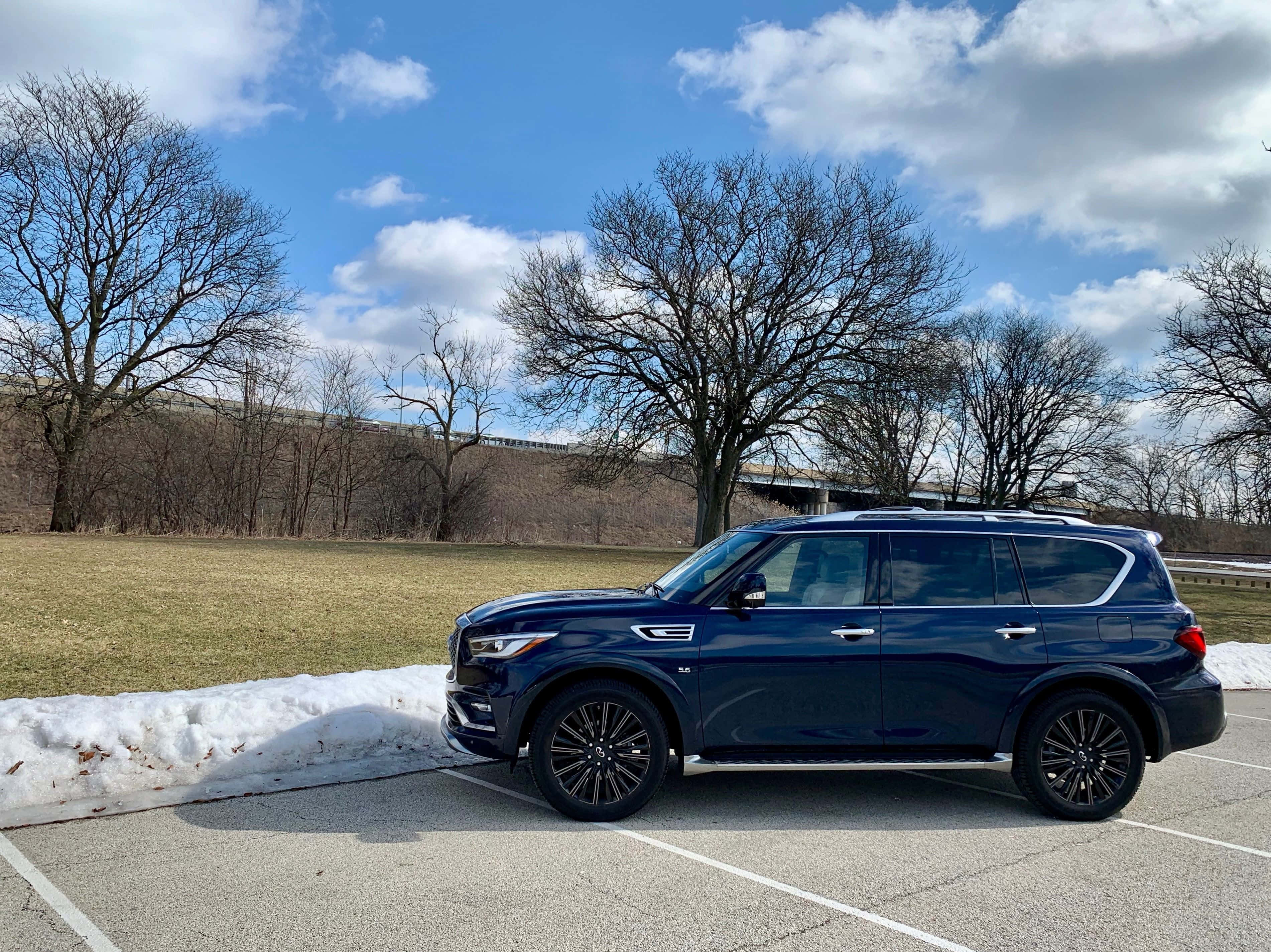 Infiniti Qx80 桌布 壁紙 — 一輛時尚的藍色SUV停在雪地停車場，上方是明亮的藍天，散落著雲朵和光禿禿的樹木。