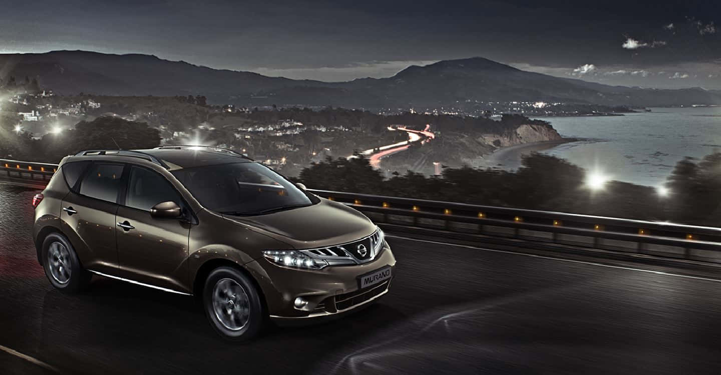 Nissan Murano 桌布 壁紙 — 一輛時尚的青銅色SUV在夜間沿著海岸公路行駛，背景是城市燈光和山脈。