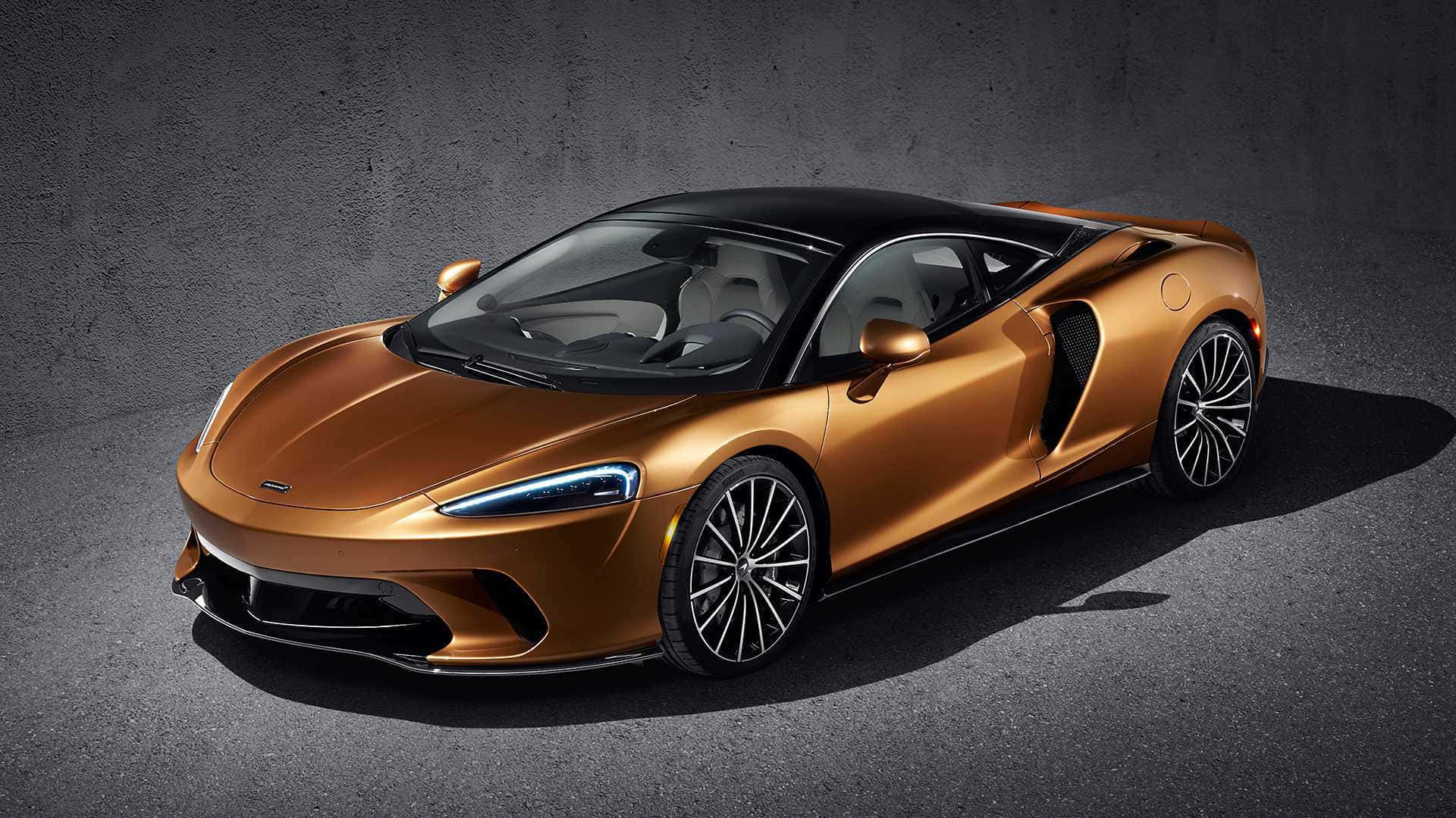 Mclaren Gt 桌布 壁紙 — 一輛時尚的銅色McLaren跑車，帶有黑色飾邊和發光的頭燈，停在深色紋理地板上，背景是灰色混凝土牆。