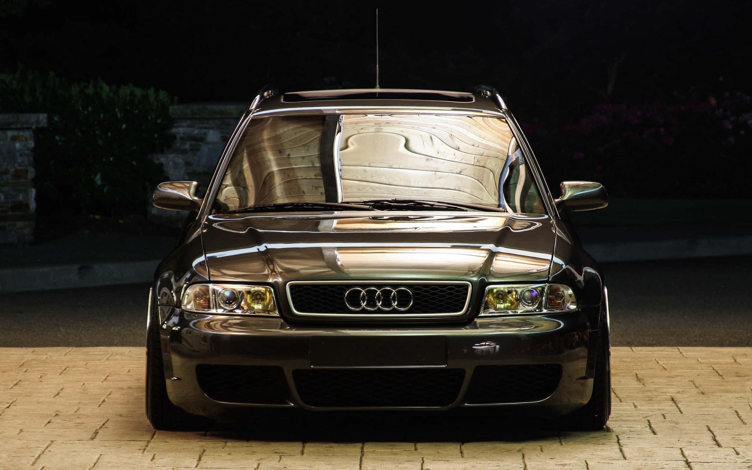 Audi S4 桌布 壁紙 — 一輛時尚的深色奧迪旅行車在夜間車道上聚光燈下閃耀，背景是模糊的樹葉。