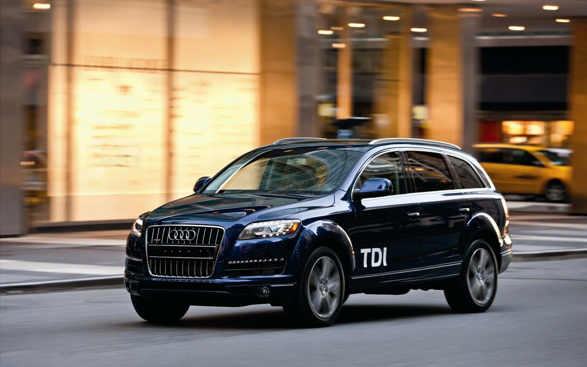 Audi Q7 桌布 壁紙 — 一輛時尚的深藍色Audi Q7 TDI SUV在城市街道上行駛，背景是模糊的商店櫥窗和一輛黃色計程車。