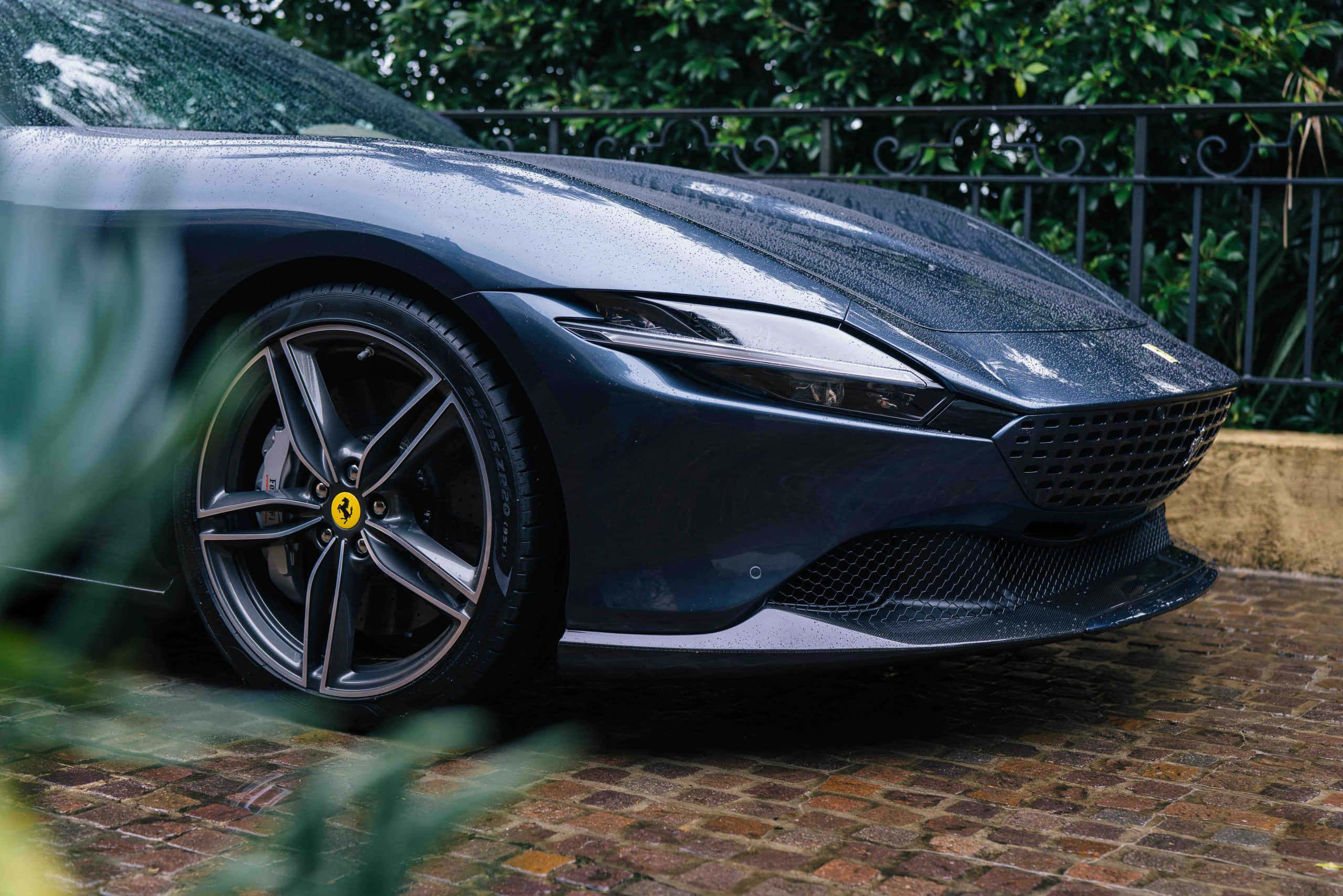 Ferrari Roma 桌布 壁紙 — 一輛時尚深藍色法拉利跑車閃爍著雨滴，停泊在由茂密綠植環繞的鵝卵石車道上。