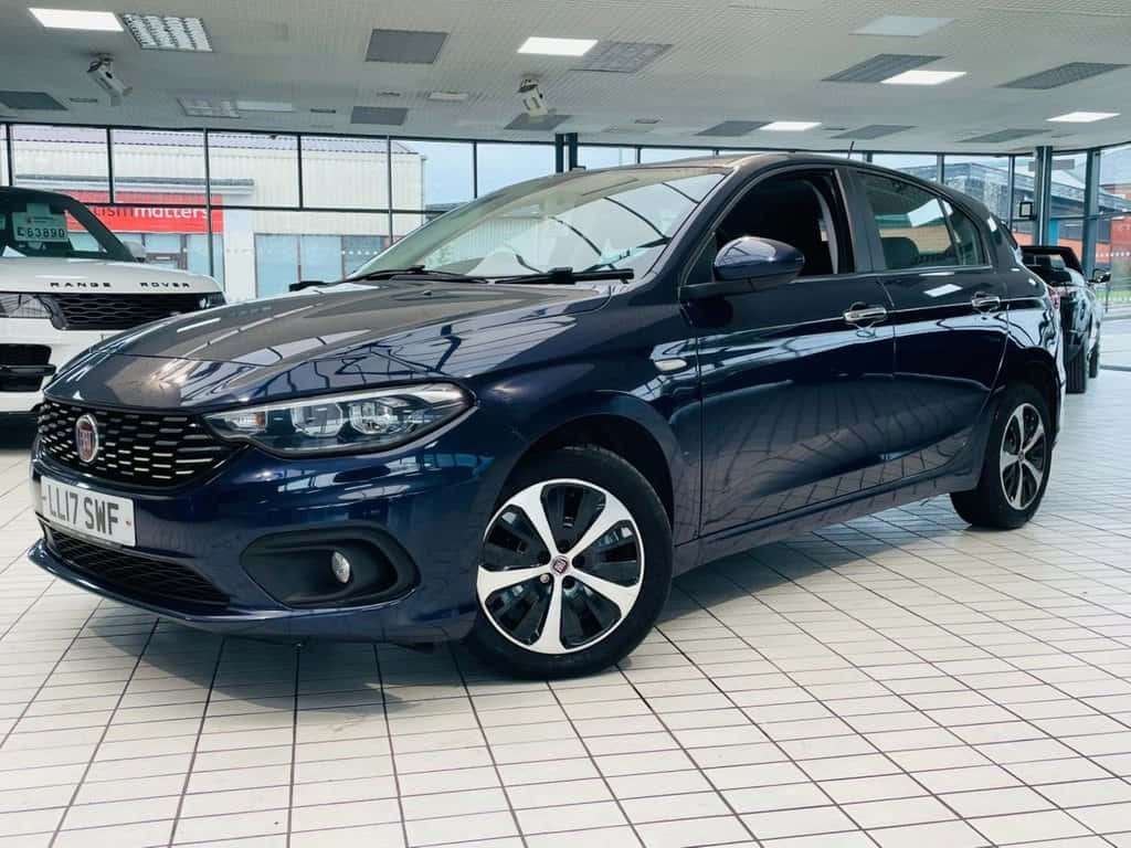 Fiat Tipo 桌布 壁紙 — 一輛時尚深藍色Fiat Punto掀背車在展示間燈光下閃閃發光，地板鋪設磁磚，背景可見其他車輛。