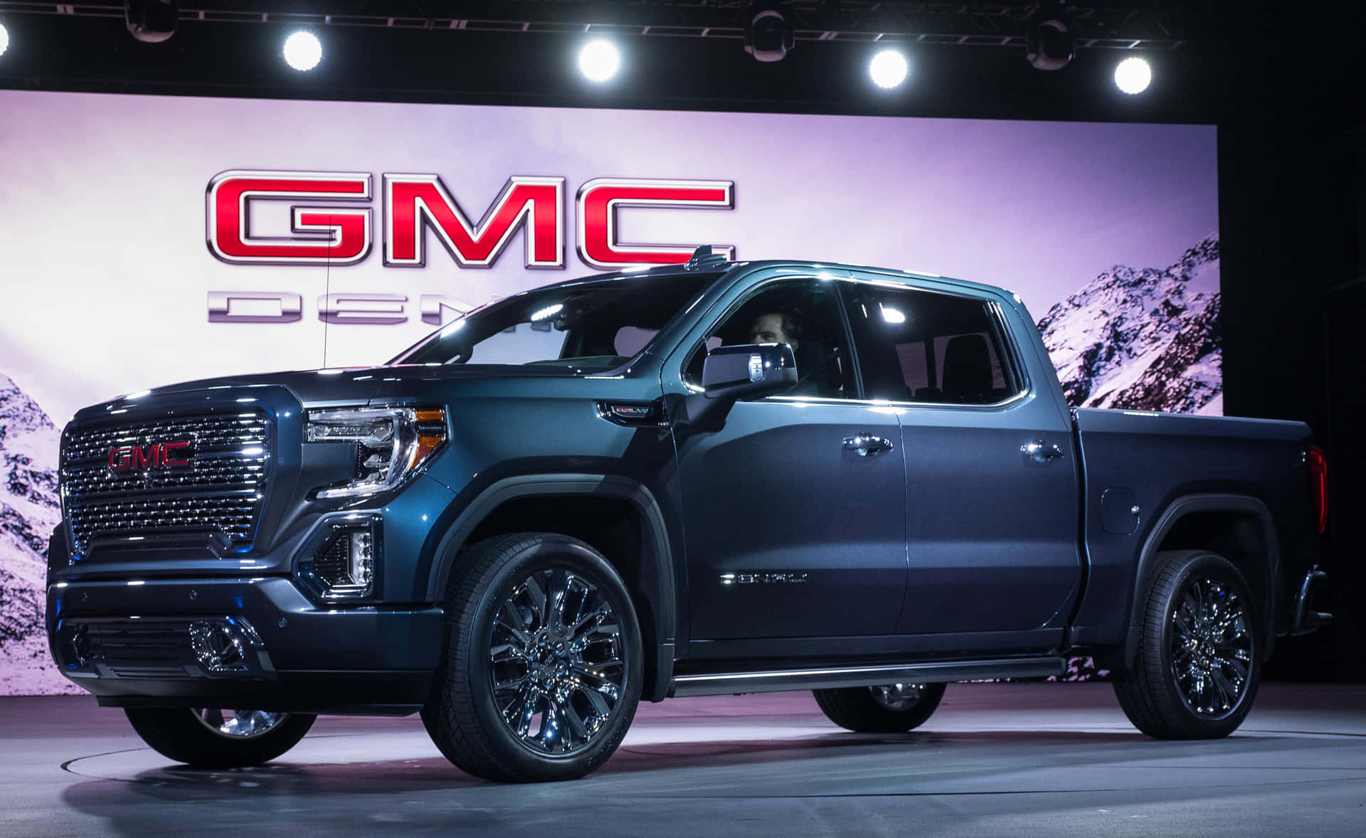 Gmc Sierra 桌布 壁紙 — 一輛時尚的深藍色 GMC Sierra Denali 皮卡車在明亮的聚光燈下於舞台上展示，背景為山脈。