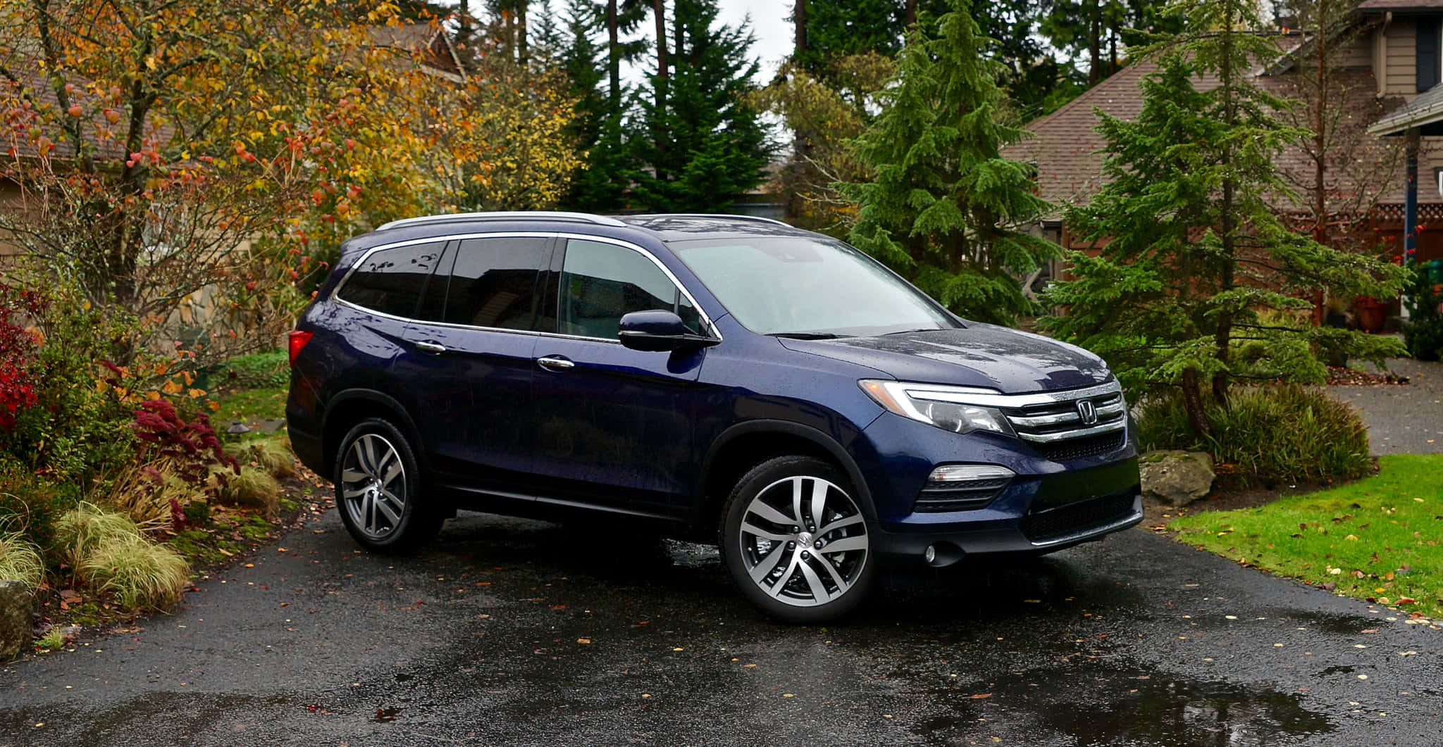 Honda Pilot 桌布 壁紙 — 一輛時尚的深藍色本田Pilot SUV停放在濕漉漉的車道上，周圍環繞著秋葉和常綠樹木。