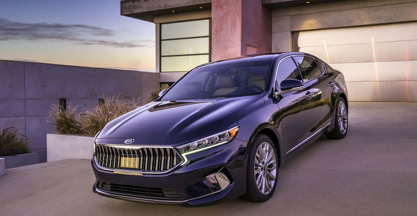 Kia Cadenza 桌布 壁紙 — 一輛時尚的深藍色Kia轎車在暮色中閃耀，停在一棟帶有溫暖車庫照明的現代混凝土房屋前。