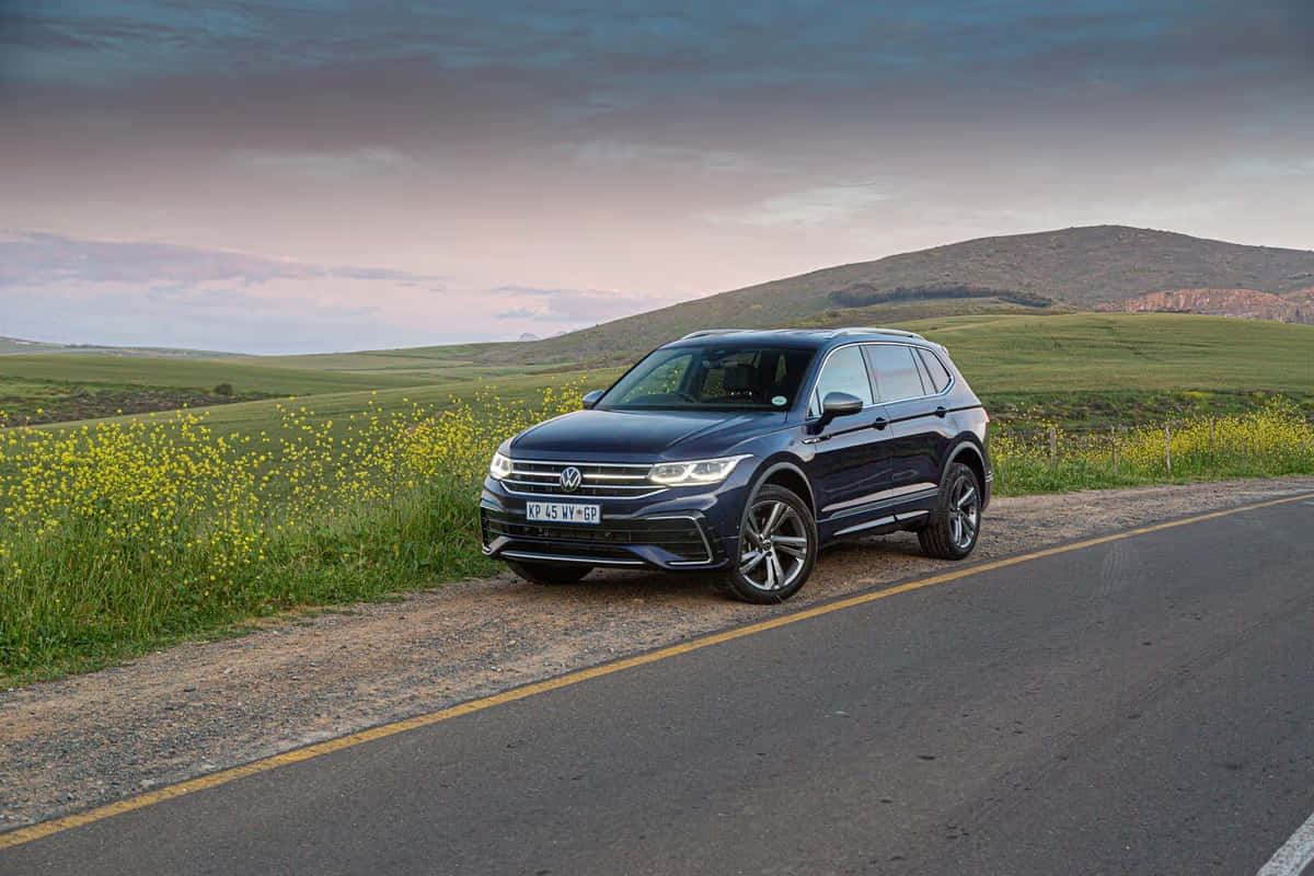 Volkswagen Tiguan 桌布 壁紙 — 一輛時尚的深藍色SUV停泊在鄉村道路上，旁邊是鮮豔的黃色野花，背景是陰鬱的黃昏天空。