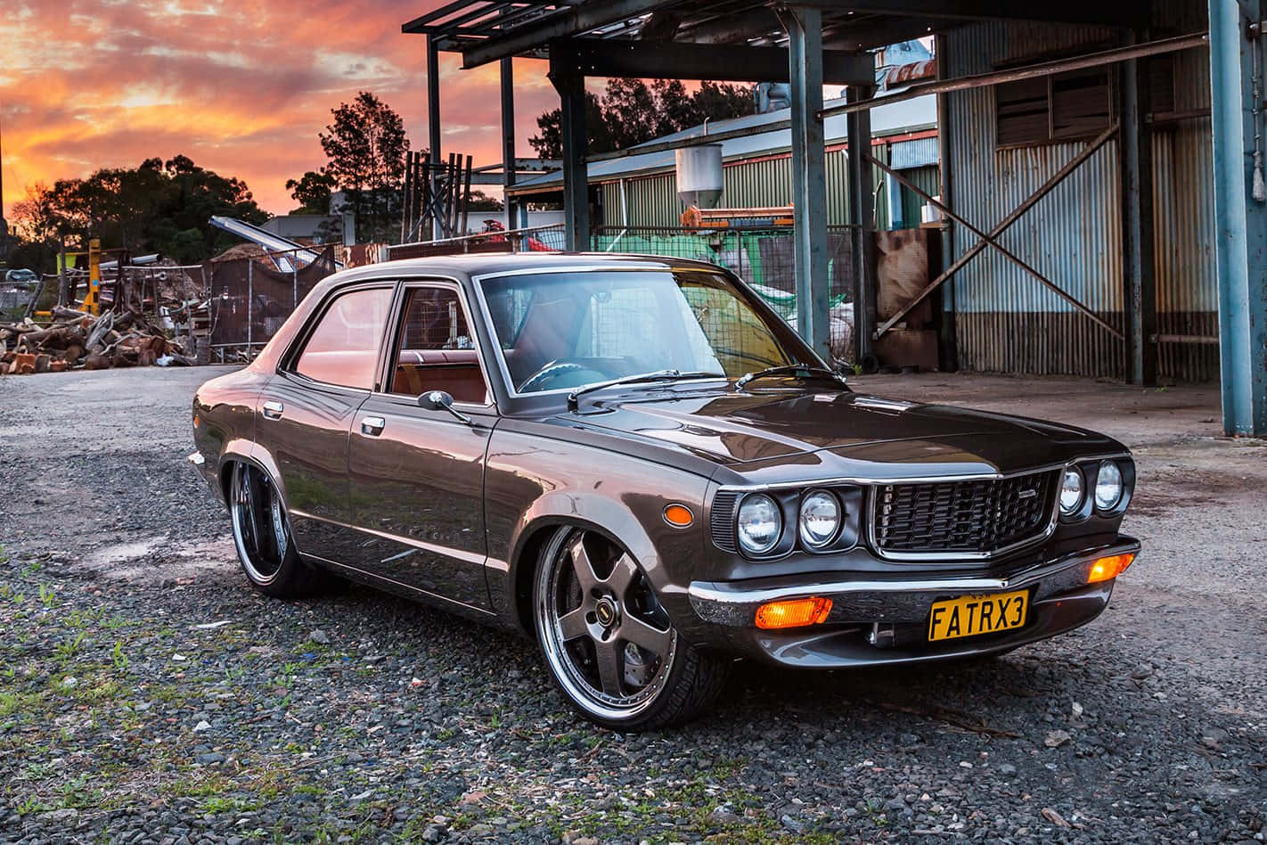 Mazda Rx-3 桌布 壁紙 — 一輛時尚深棕色經典汽車配備定制輪圈，停在夕陽下生鏽工業建築前的碎石地上。