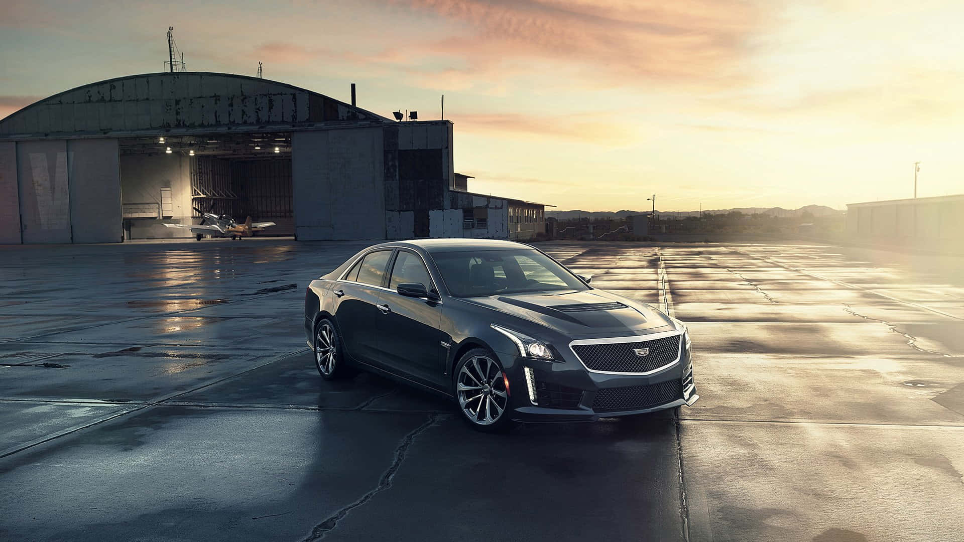 Cadillac Cts 桌布 壁紙 — 一輛時尚的深灰色 Cadillac CT5-V 停在黃金時段開放機庫前的濕滑停機坪上。