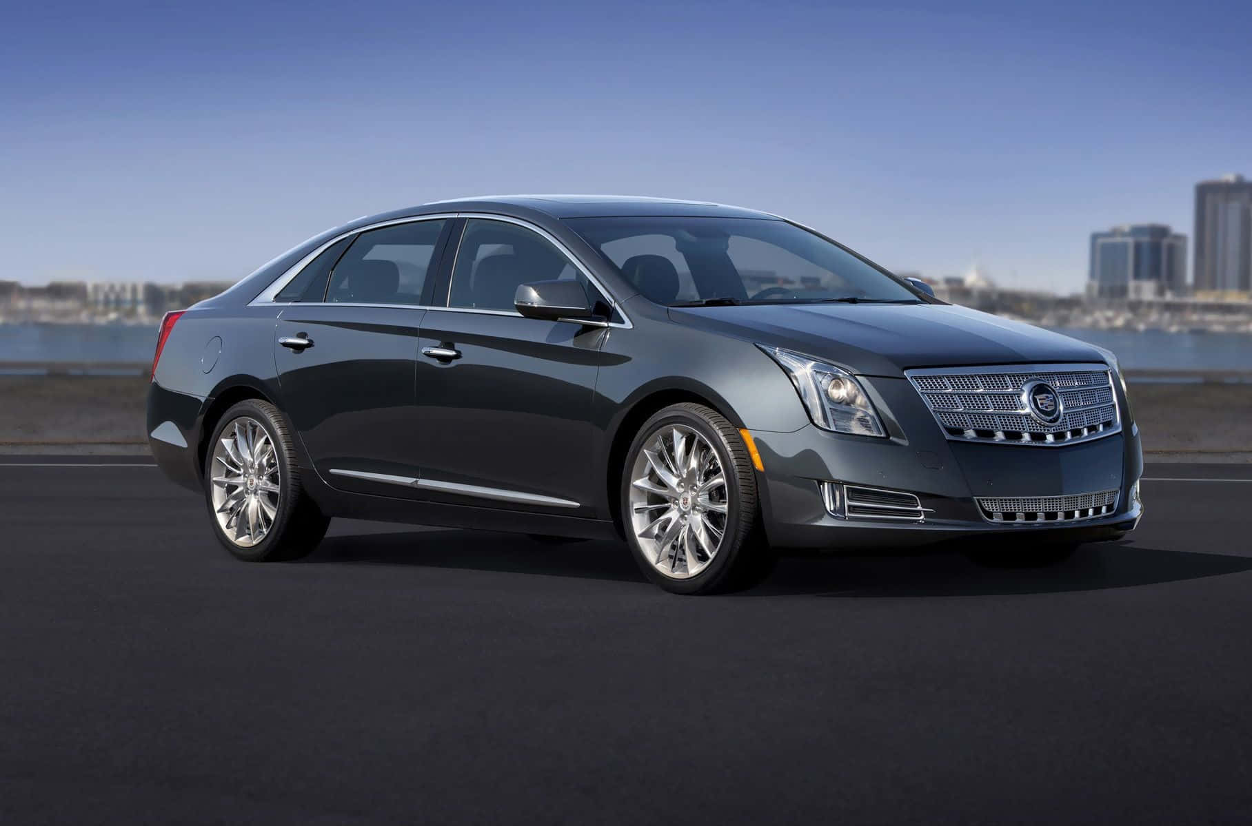 Cadillac Xts 桌布 壁紙 — 一輛時尚的深灰色Cadillac轎車停在海濱道路上，背景是城市天際線，天空湛藍清澈。
