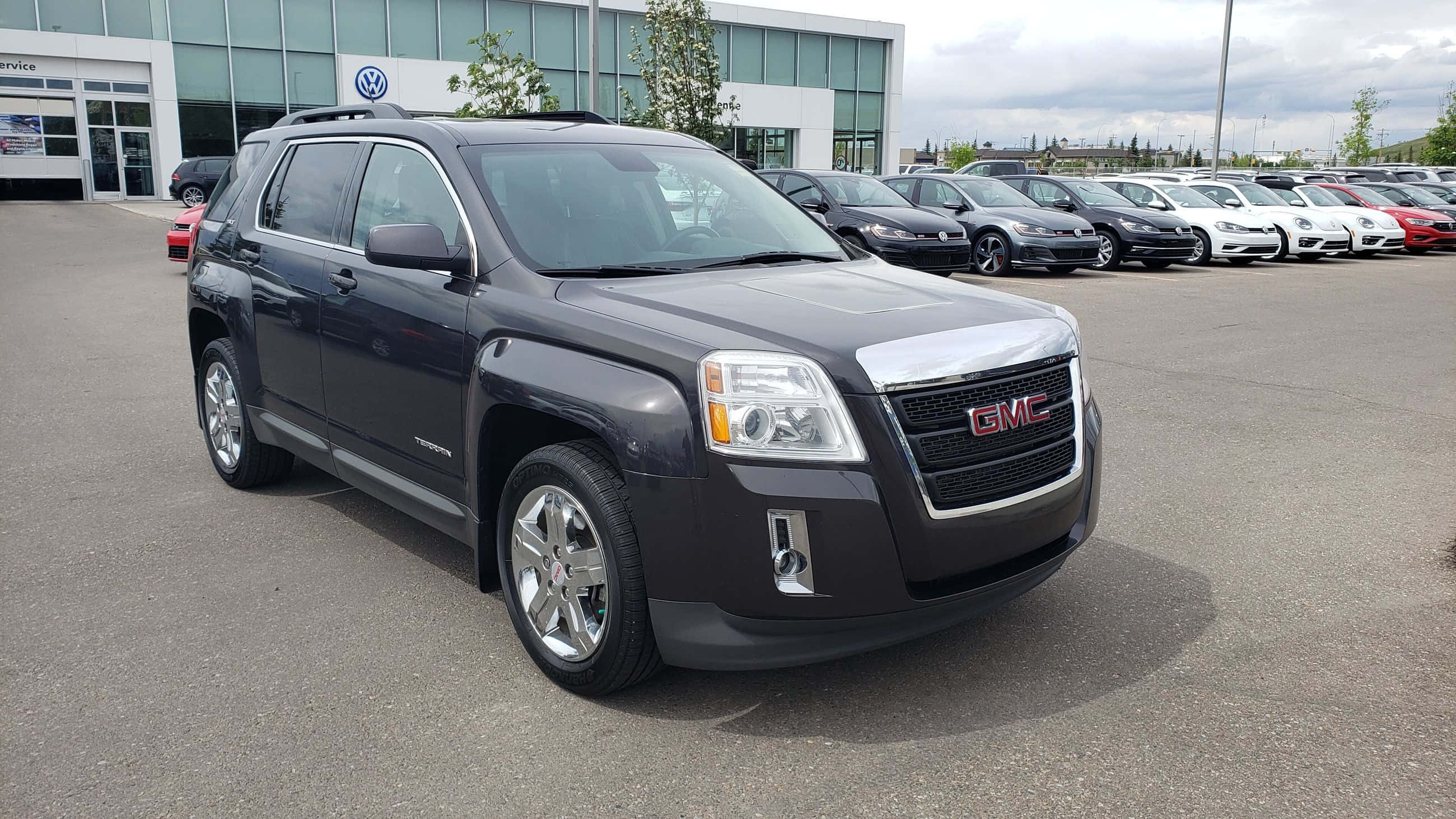 Gmc Terrain 桌布 壁紙 — 一輛時尚的深灰色 GMC Terrain SUV 停放在經銷商場地，與其他車輛一起，天空多雲。