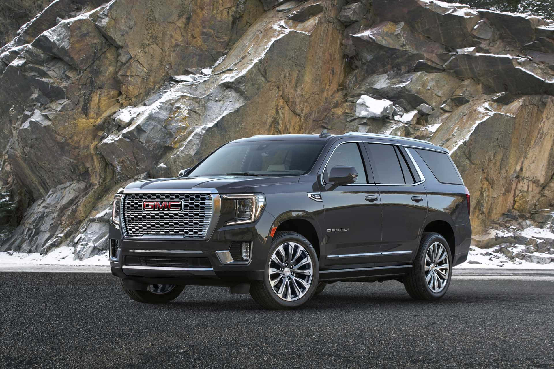 Gmc Yukon 桌布 壁紙 — 一輛時尚深灰色GMC Yukon Denali SUV停泊在瀝青路上，前方是崎嶇積雪的岩石懸崖面。