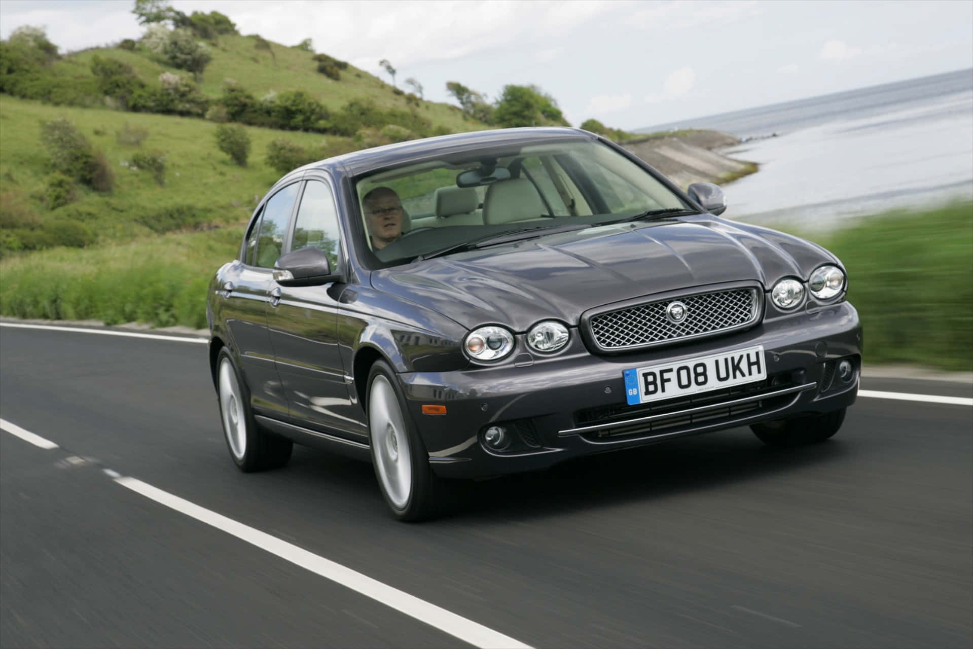 Jaguar X-type 桌布 壁紙 — 一輛時尚的深灰色積架轎車沿著海岸公路疾馳，背景是綠色山丘和海洋。