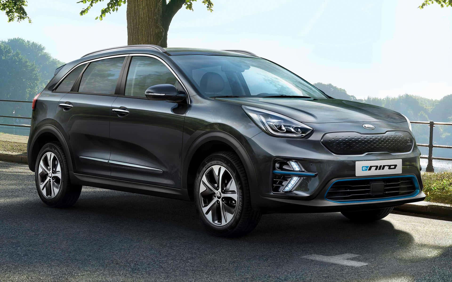 Kia Niro 桌布 壁紙 — 一輛時尚深灰色Kia e-Niro電動SUV停泊在鋪砌道路上，旁邊有樹木，背景是風景如畫的湖泊和森林覆蓋的山丘。