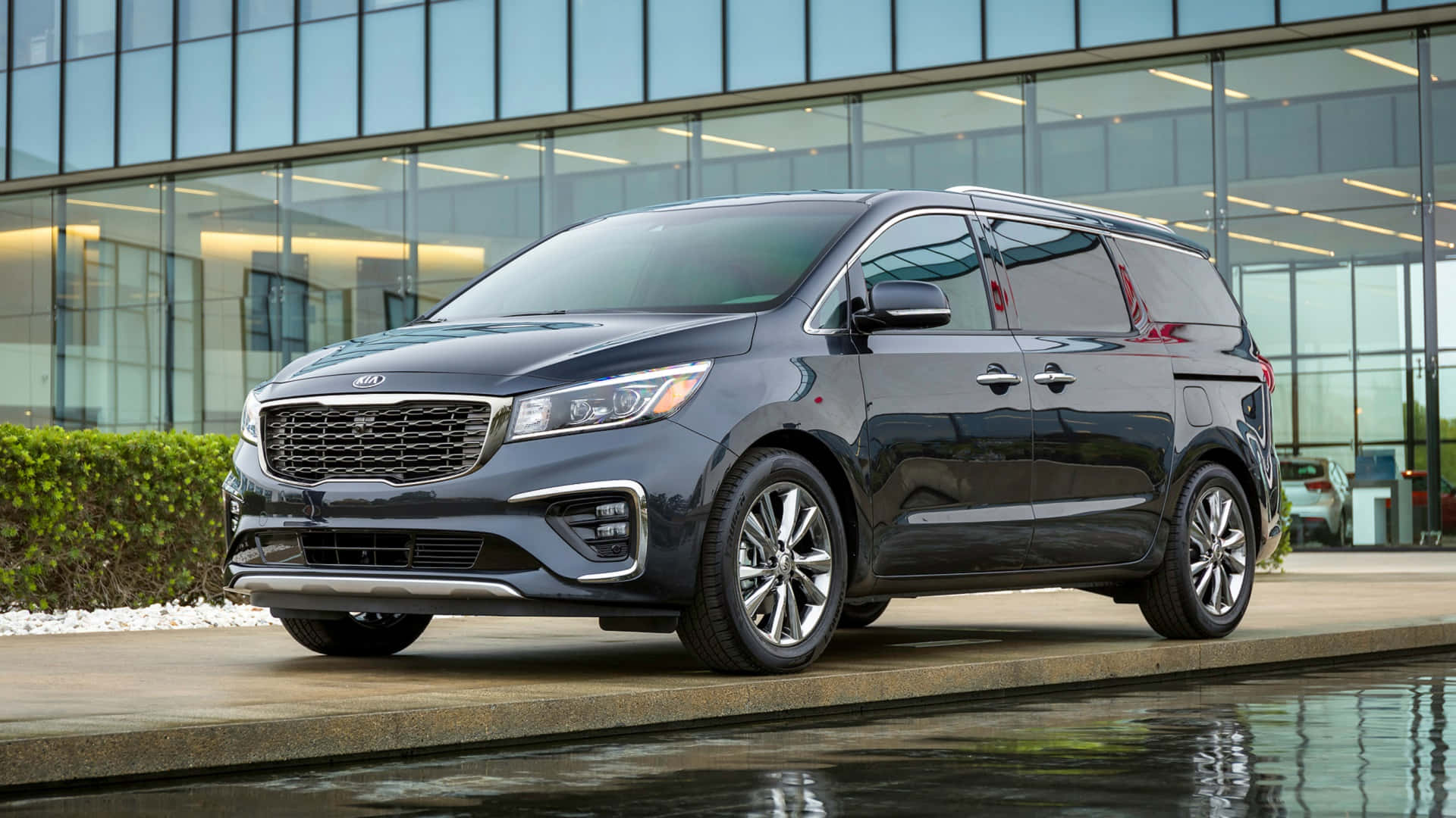 Kia Sedona 桌布 壁紙 — 一輛時尚深灰色Kia廂型車停放在現代玻璃建築前的反光水景旁邊。