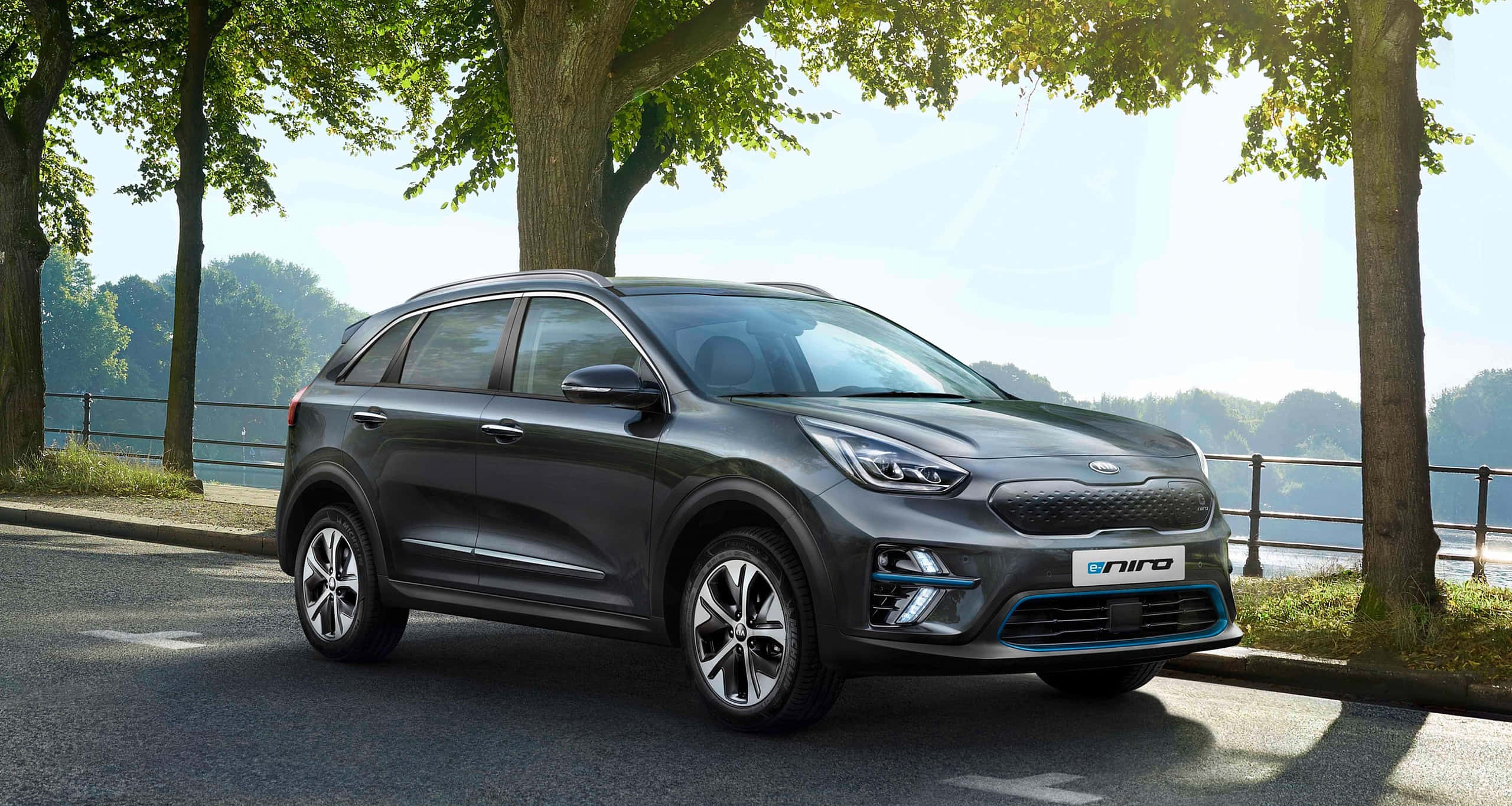 Kia Niro 桌布 壁紙 — 一輛時尚的深灰色Kia Niro電動SUV停在陽光明媚、綠樹成蔭的湖邊道路上。