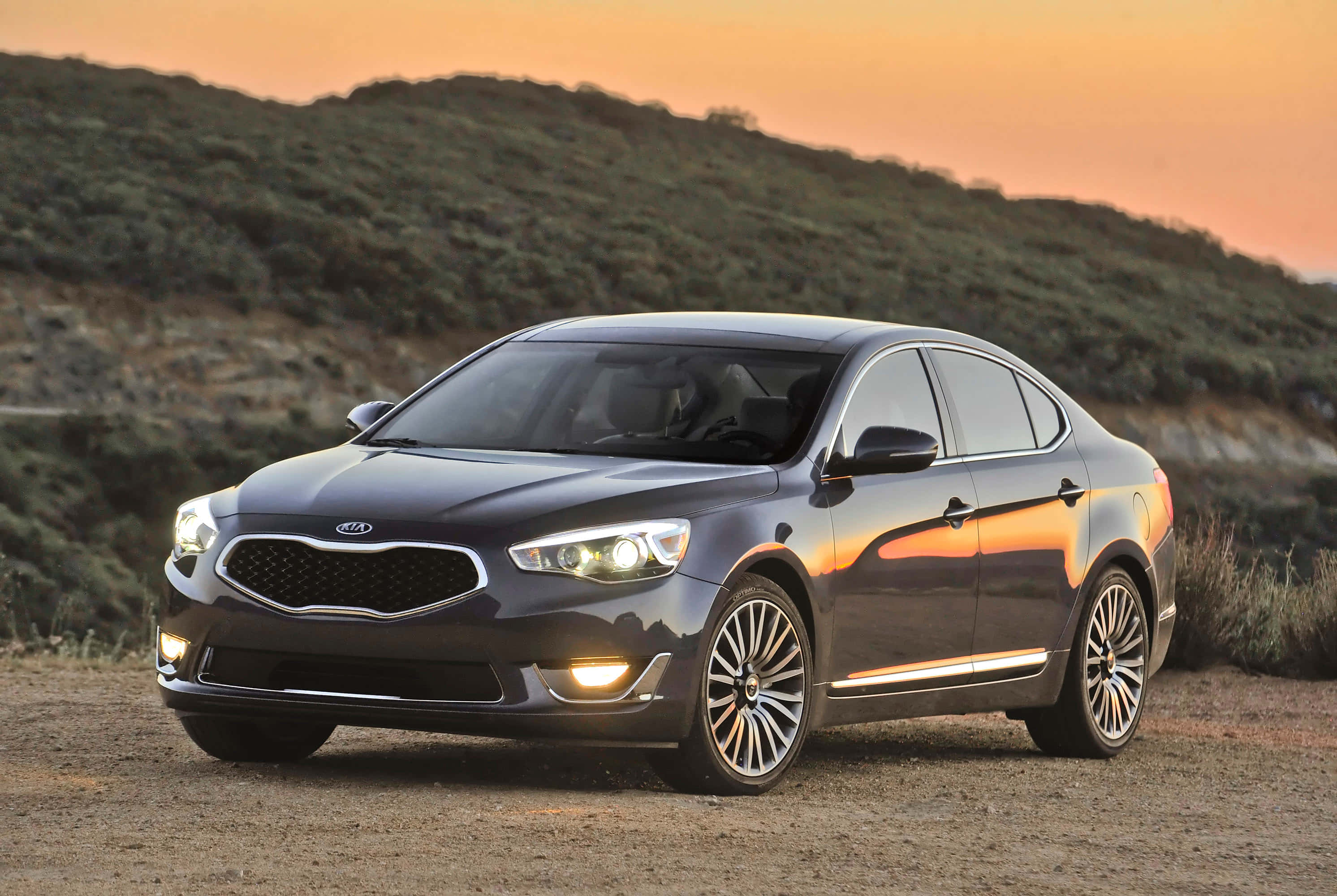 Kia Cadenza 桌布 壁紙 — 一輛流線型深灰色起亞轎車在溫暖的夕陽光下閃閃發光，停在塵土飛揚的山坡上，背景是連綿起伏的綠色山丘。