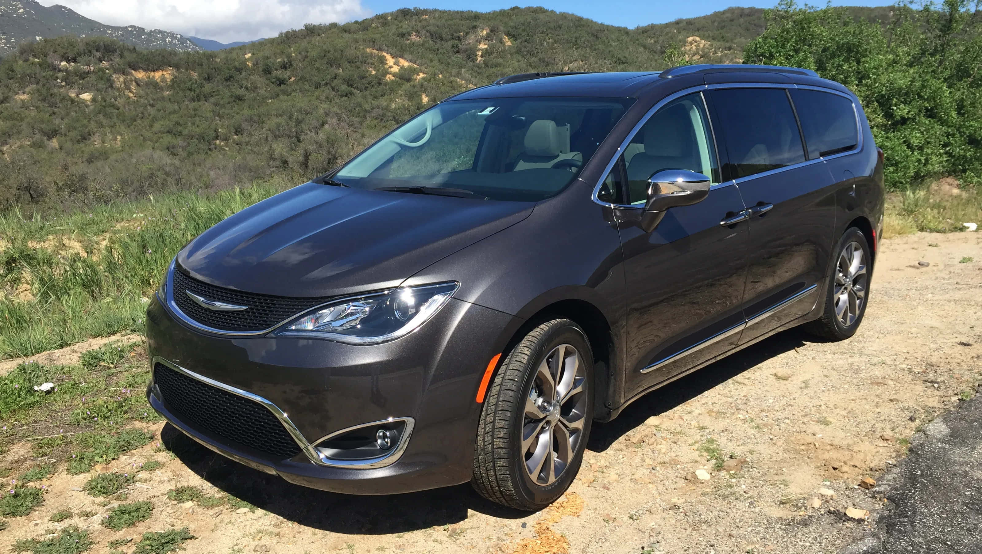 Chrysler Pacifica 桌布 壁紙 — 一輛時尚深灰色小型客車停泊在泥路上，周圍是起伏的綠色山丘和散落的樹木，頭頂是明亮的藍天。
