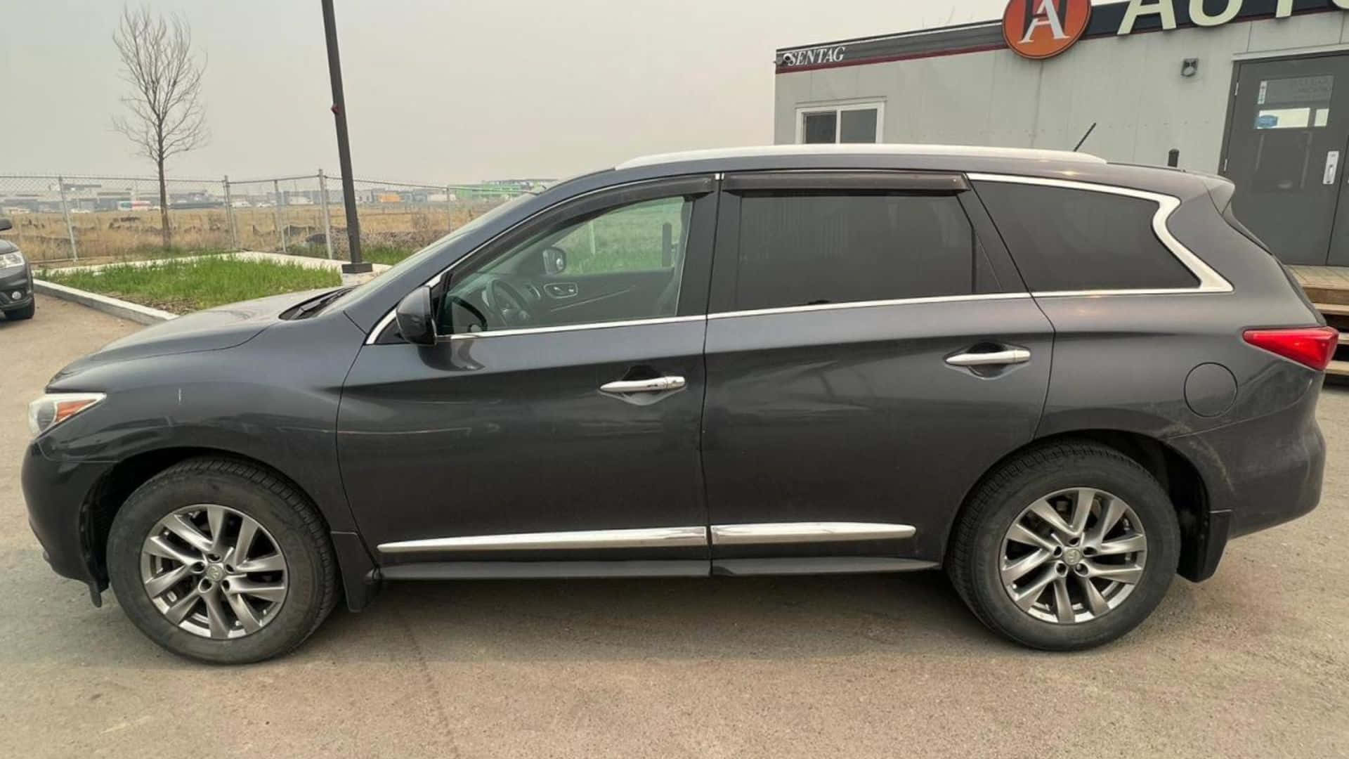 Infiniti Jx35 桌布 壁紙 — 一輛時尚的深灰色SUV停放在經銷商外面，背景有光禿的樹木和陰天。