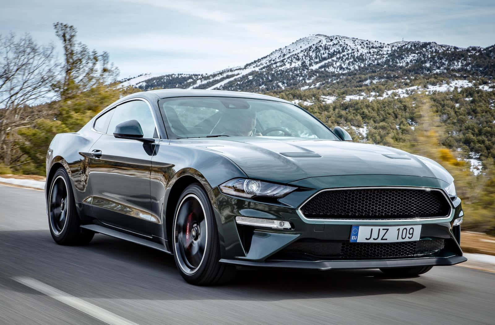 Ford Mustang Bullitt 桌布 壁紙 — 一輛流線型深綠色福特野馬在雪山頂和秋樹背景下沿山路飛馳。