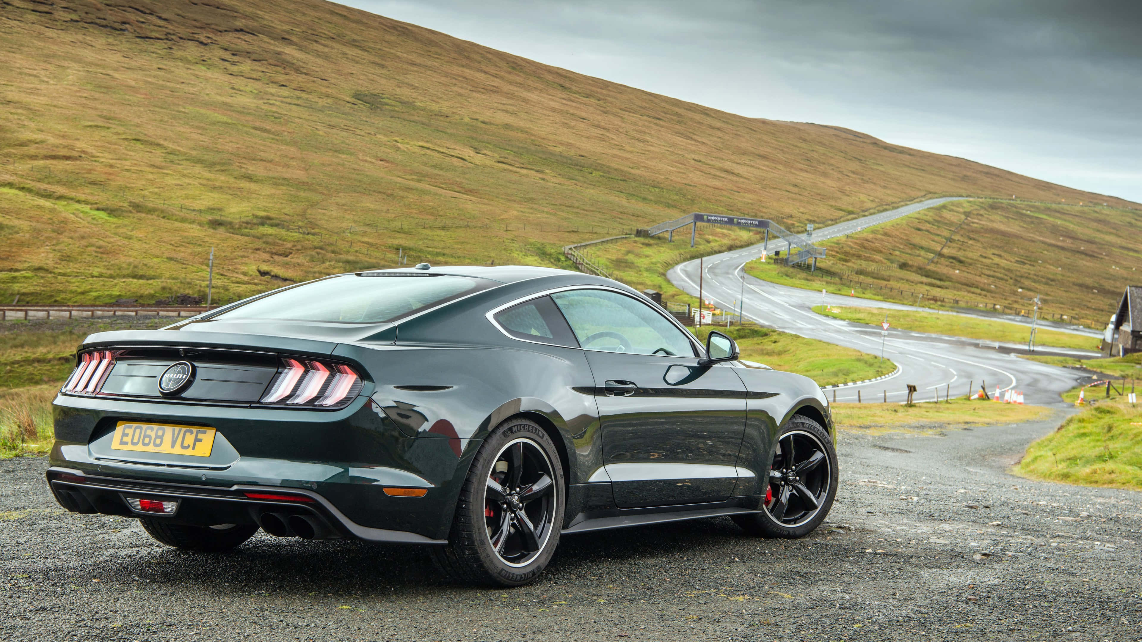 Ford Mustang Bullitt 桌布 壁紙 — 一輛流線型深綠色福特野馬停泊在蜿蜒穿過草坡的碎石路上，天空多雲。