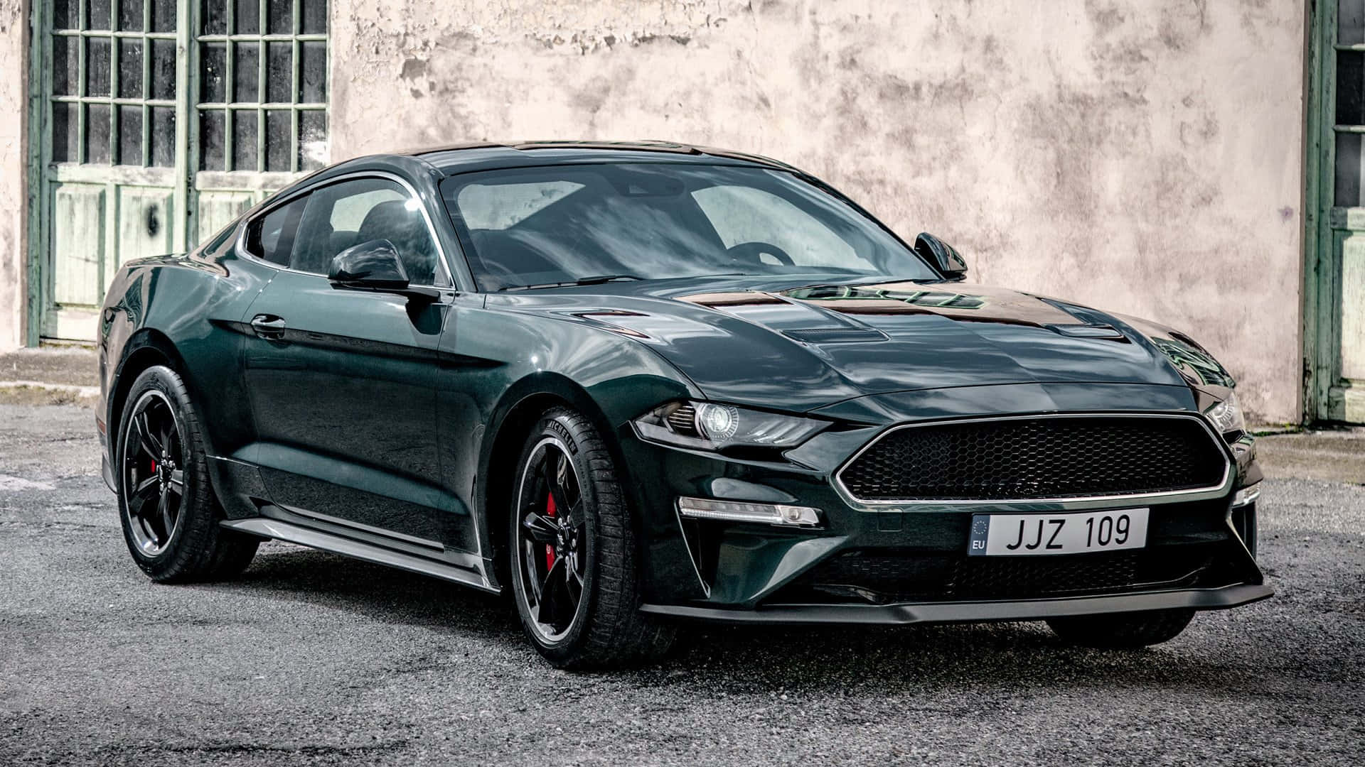 Ford Mustang Bullitt 桌布 壁紙 — 一輛時尚深綠色福特野馬跑車停泊在碎石路上，前方是一座風化的工業建築。