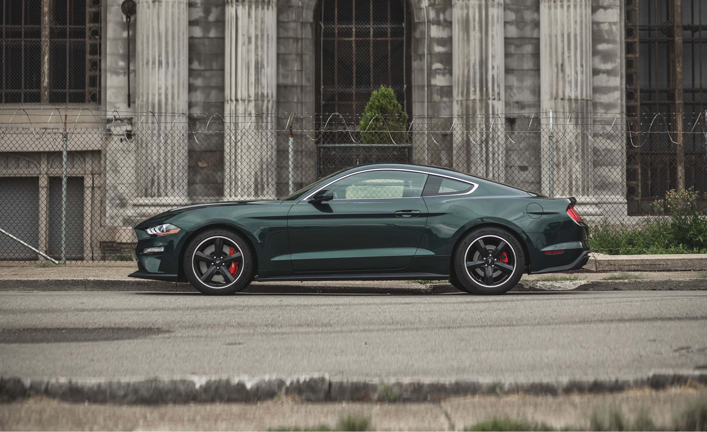 Ford Mustang Bullitt 桌布 壁紙 — 一架時尚的深綠色福特野馬跑車停放在佈滿灰塵的城市街道上，旁邊是鐵絲網圍欄和風化的石建築。