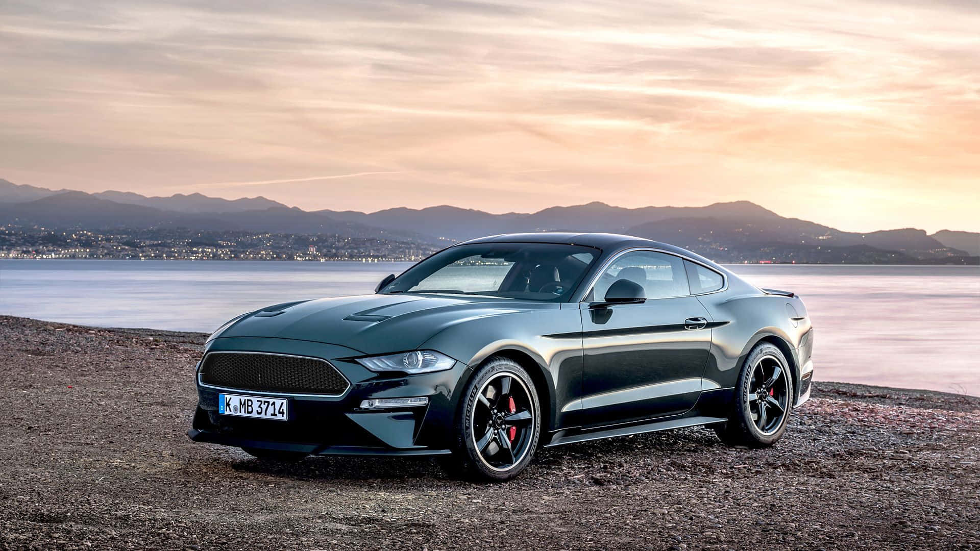 Ford Mustang Bullitt 桌布 壁紙 — 一輛時尚的深綠色福特野馬跑車停泊在日落時分的岩石海岸上，背景有山脈和水面。