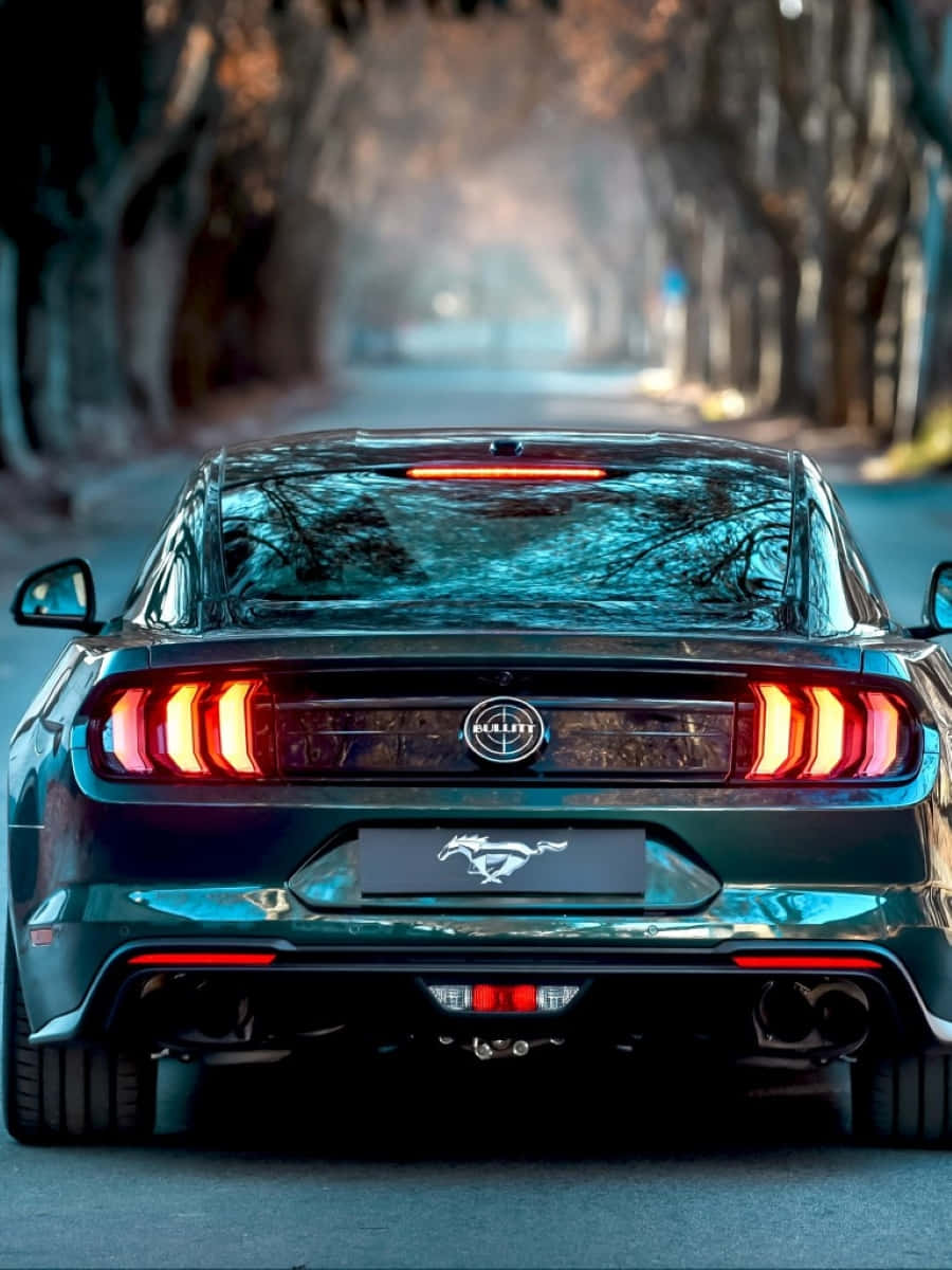 Ford Mustang Bullitt 桌布 壁紙 — 一輛時尚的深綠色野馬，帶有發光的紅色尾燈，在黃昏時分行駛在一條霧氣瀰漫的樹林道路上。