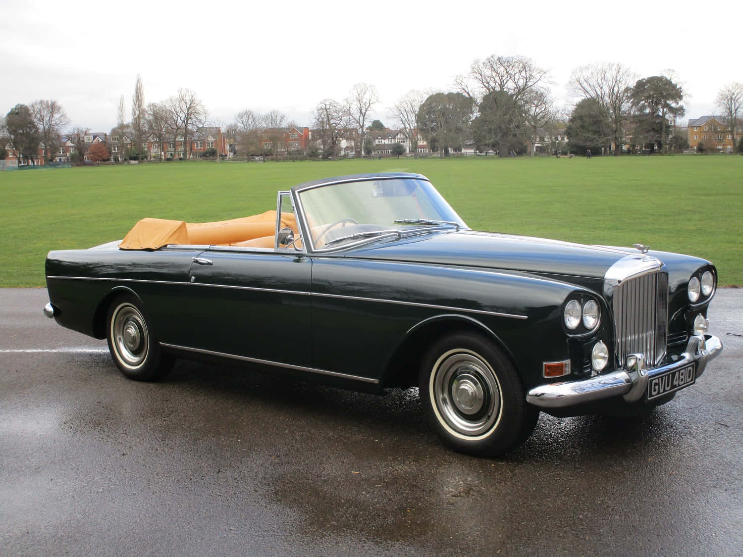 Bentley S3 桌布 壁紙 — 一輛時尚深綠色復古敞篷車，配米色內裝，停放在潮濕路面上，旁邊是廣闊的綠色田野，天空多雲。