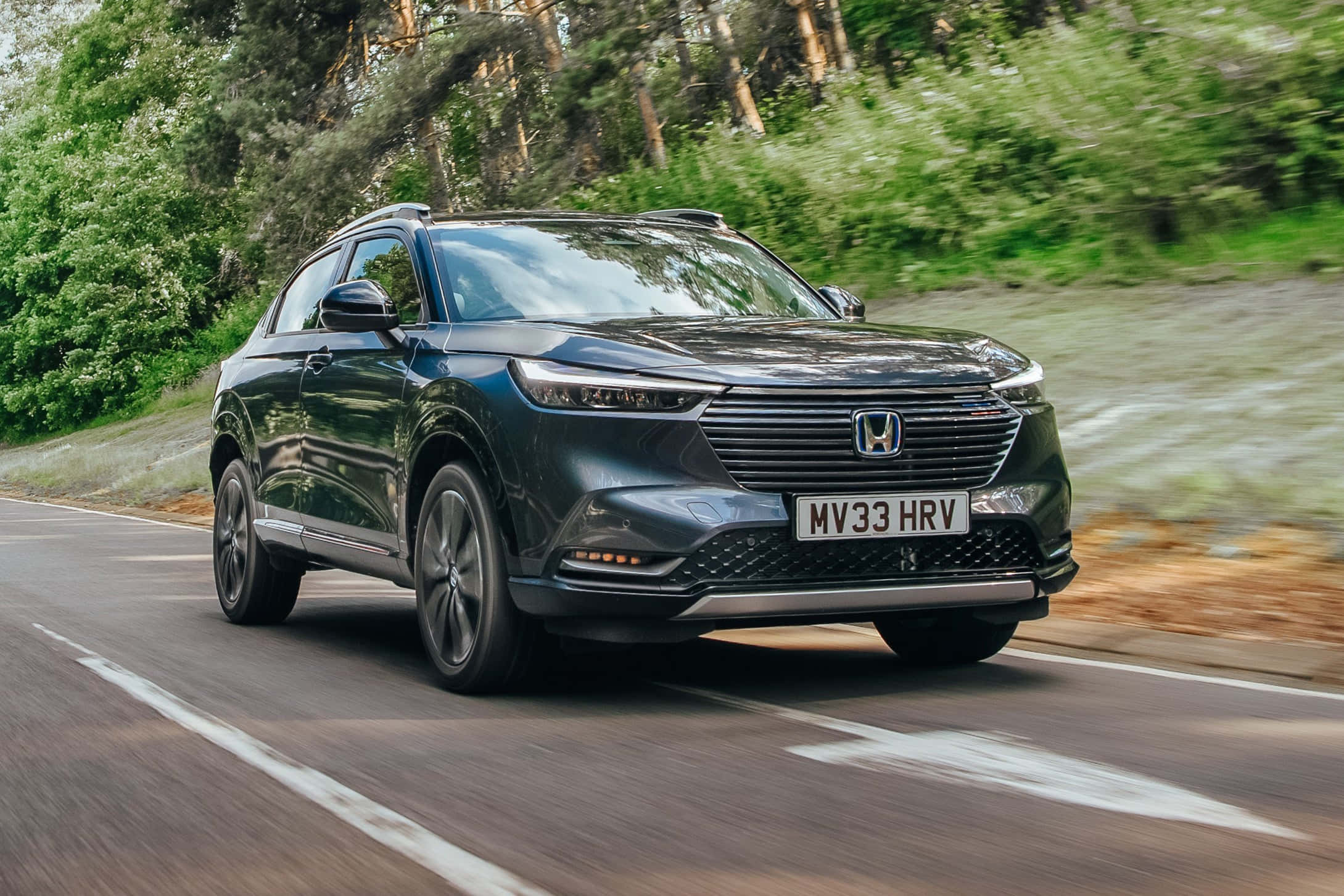 Honda Hr-v 桌布 壁紙 — 一輛時尚的深灰色本田HRV SUV沿著蜿蜒的森林道路快速行駛，周圍是模糊的綠色樹葉。