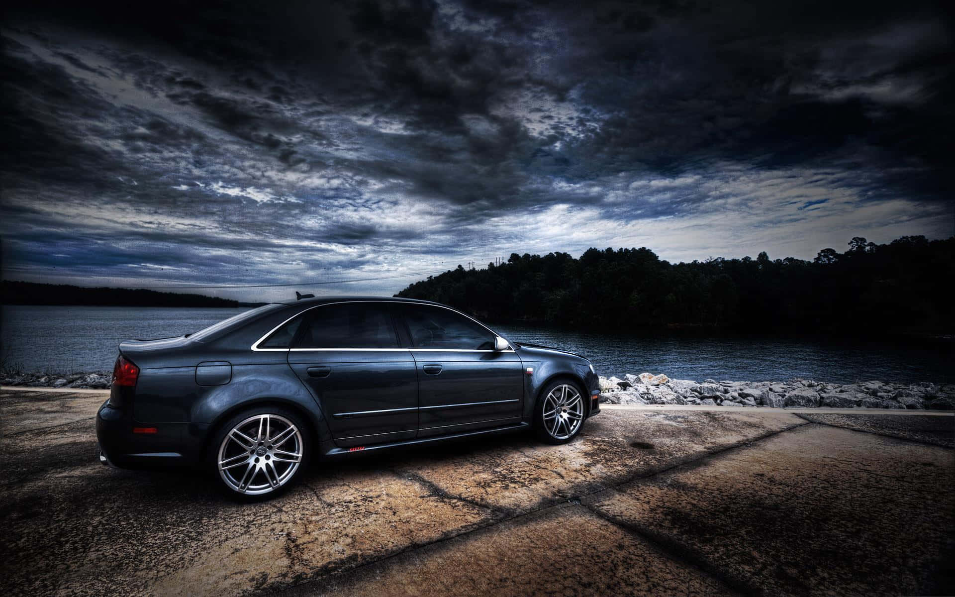 Audi Rs4 桌布 壁紙 — 一輛時尚的深色轎車停泊在裂開的混凝土岸邊，背景是戲劇性的暴風雨天空和寧靜的湖泊。