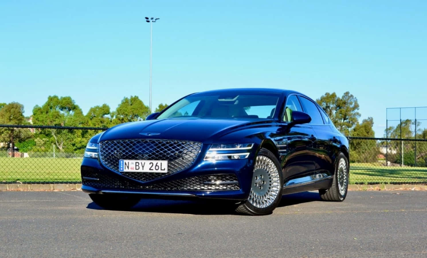 Genesis G70 桌布 壁紙 — 一輛時尚的深藍色豪華轎車在明亮的陽光下閃閃發光，停在柏油路上，背景有綠樹和圍欄。