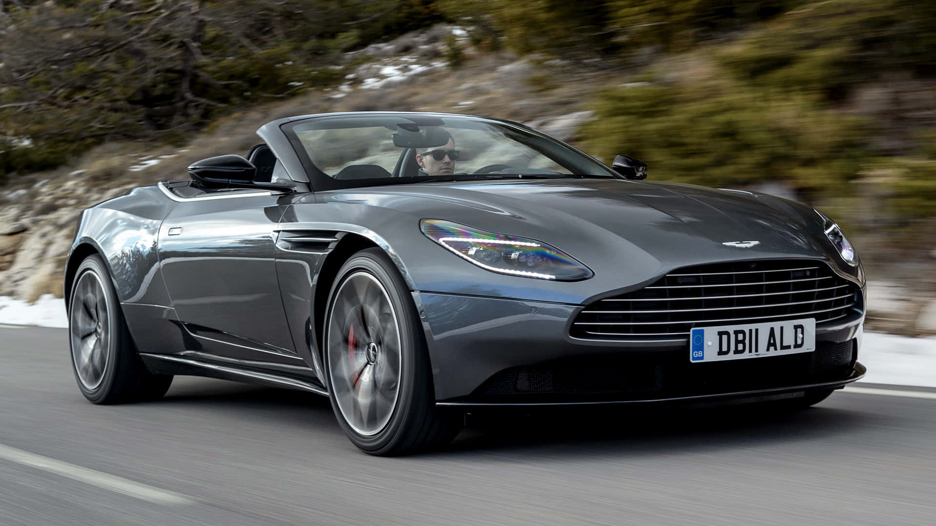 Aston Martin Db11 桌布 壁紙 — 一輛時尚的灰色 Aston Martin DB11 敞篷跑車沿著蜿蜒的山路疾馳，兩旁是白雪覆蓋的懸崖和模糊的綠色植物。