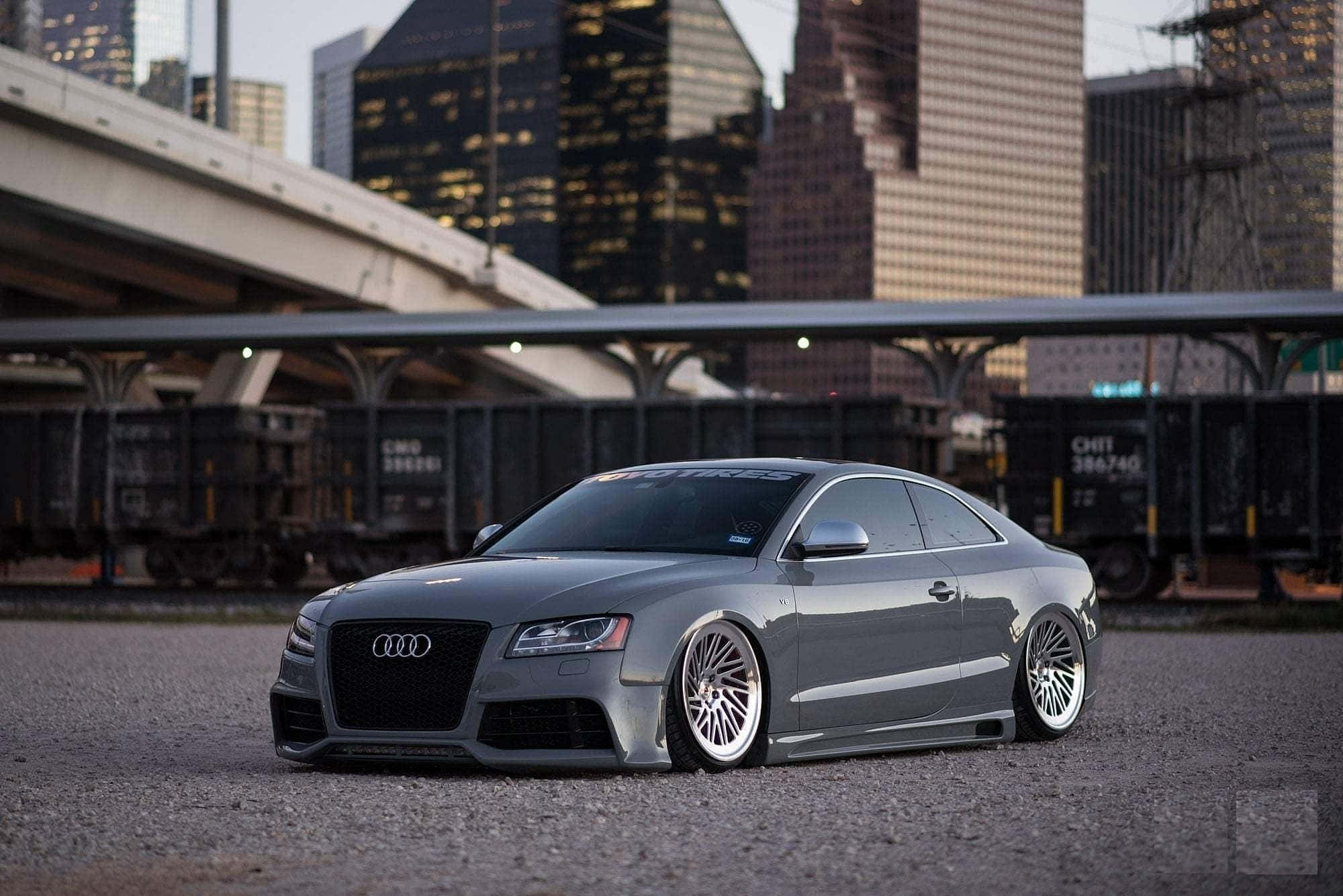 Audi S5 桌布 壁紙 — 一輛時尚的灰色奧迪雙門轎跑車配備訂製輪圈，在黃昏時分低趴於城市天橋下的碎石路上，靠近貨運列車。